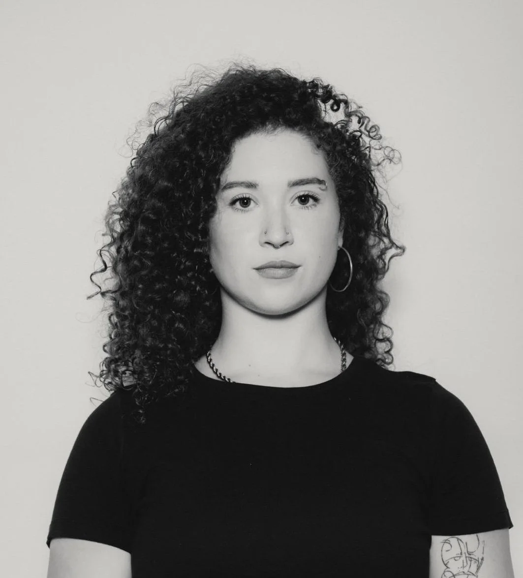 Black and white portrait of a woman with curly hair, wearing a black top, hoop earrings, and a nose ring, standing against a plain background.