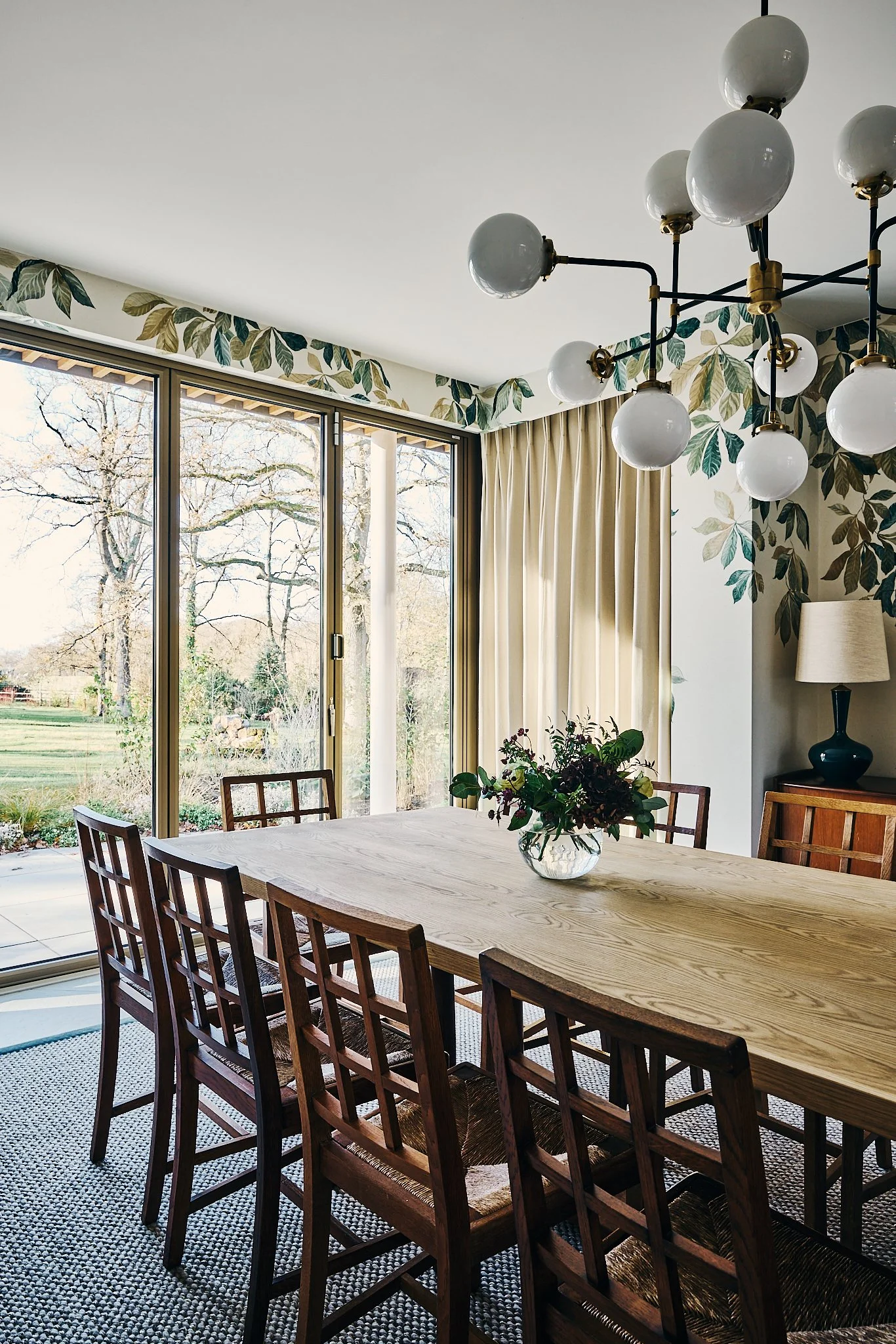 A dining room with a wooden table and six wooden chairs, a glass vase with flowers, large windows with beige curtains, a floral leaf border on the wall, a modern chandelier with multiple white spherical bulbs, and a side table with a lamp.