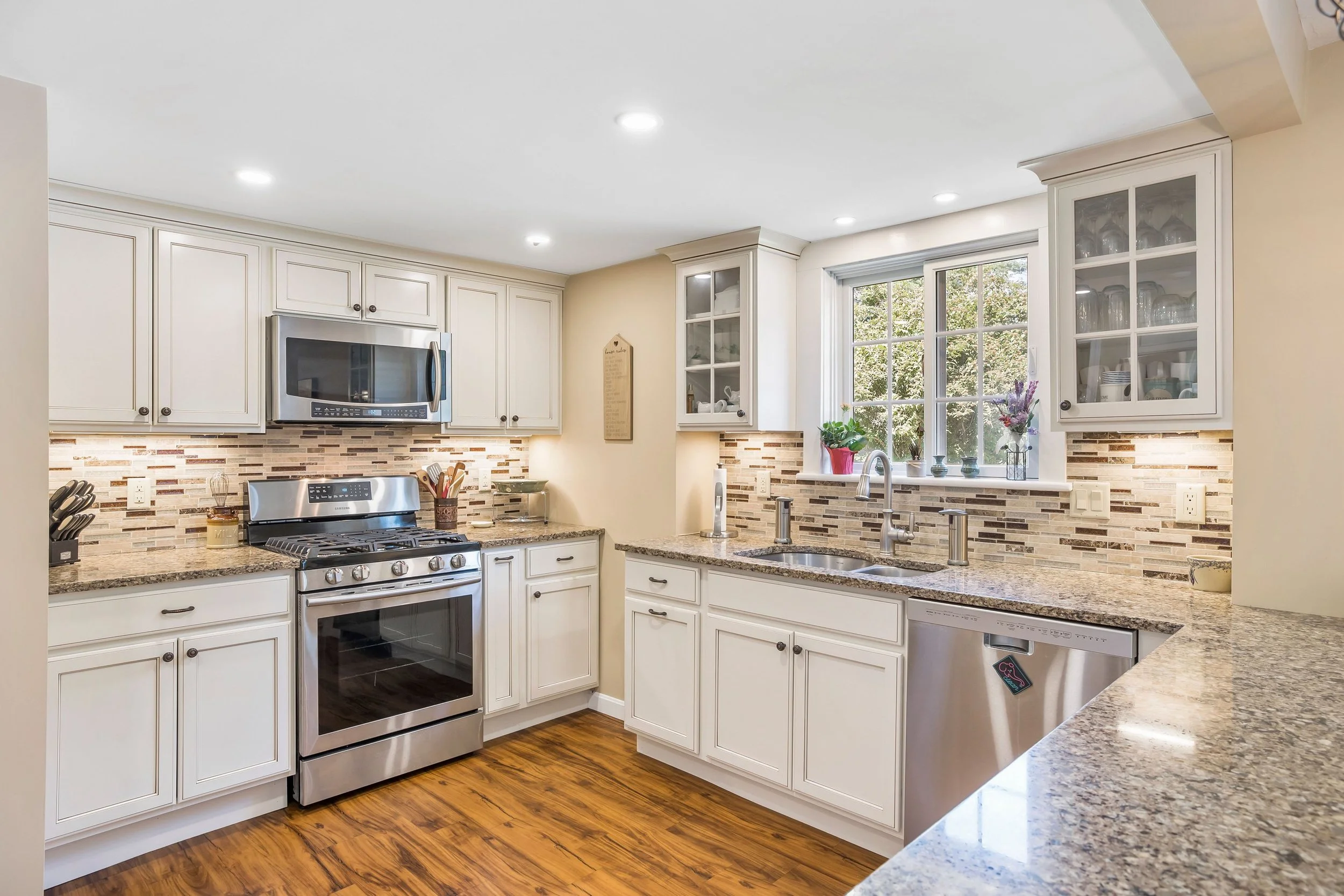 Bright kitchen with white cabinets, granite countertops, stainless steel appliances, a window above the sink, and a colorful backsplash.