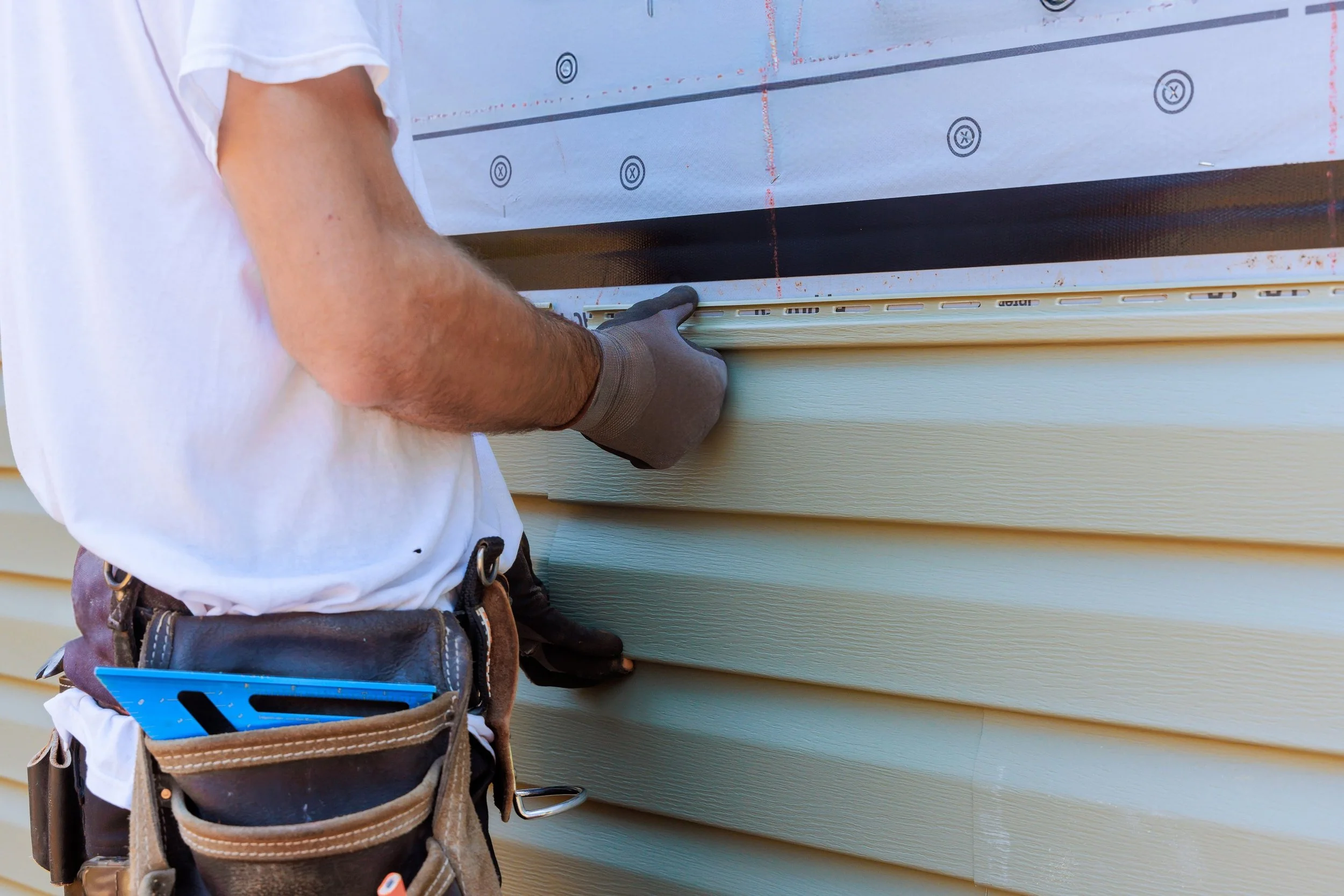 A worker installing vinyl siding on a house, wearing a tool belt and gloves, and securing siding panels to the exterior wall.