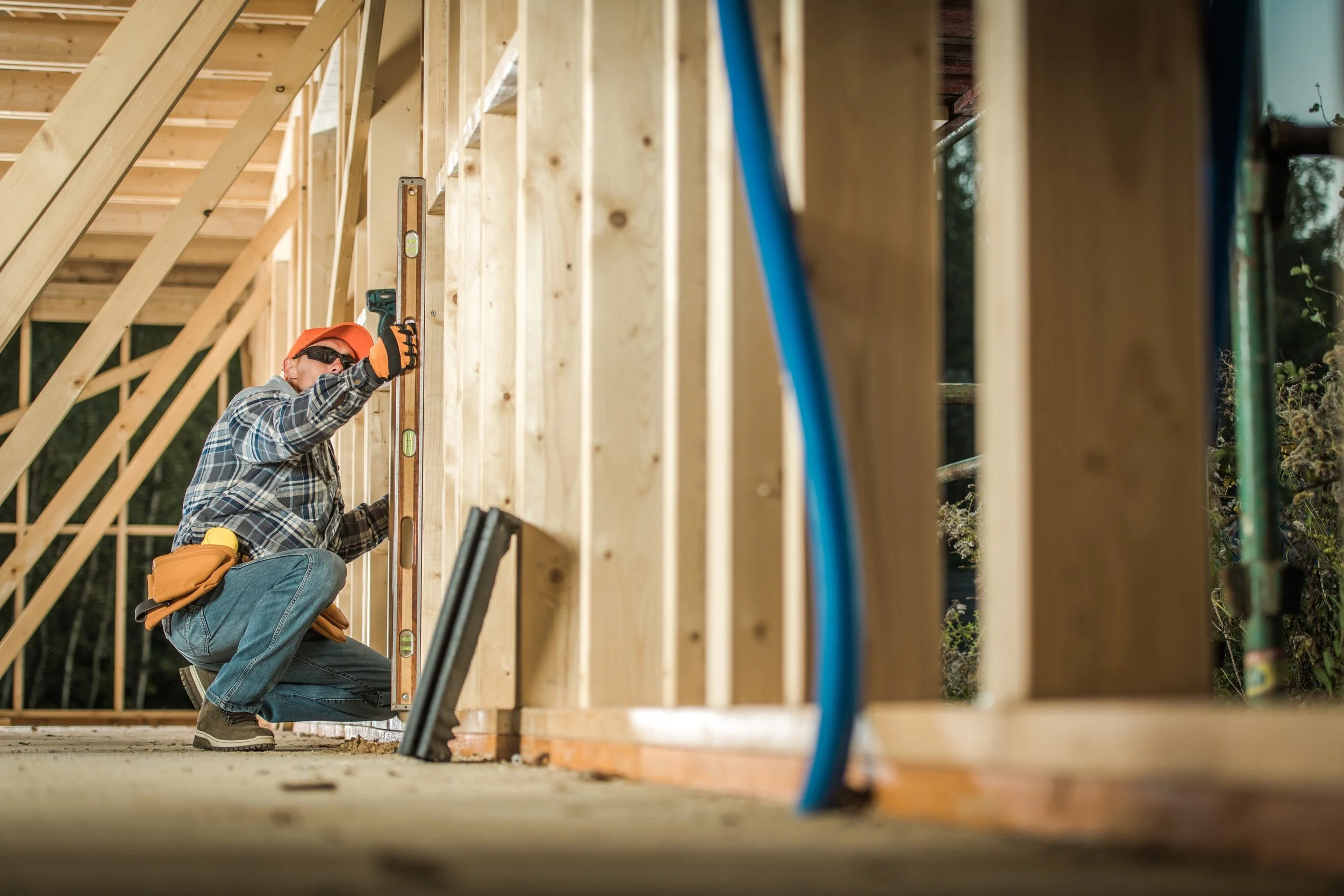A construction worker in plaid shirt, jeans, orange hat, and sunglasses kneeling and using a level on a wooden wall frame at a building site.