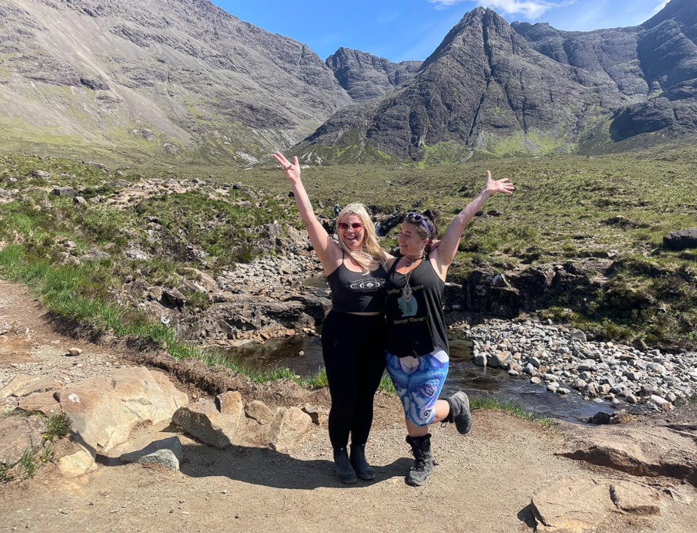 Two women smiling and celebrating with arms raised in a mountain valley, with rocky terrain, a small stream, and tall rugged mountains in the background under a partly cloudy sky.