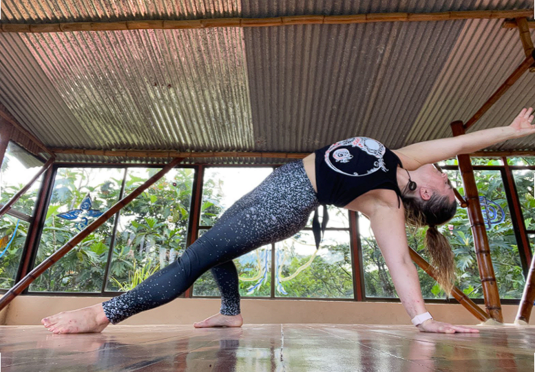 A woman practicing yoga in a studio with large windows, performing a downward dog pose with her left foot forward and right leg extended back.