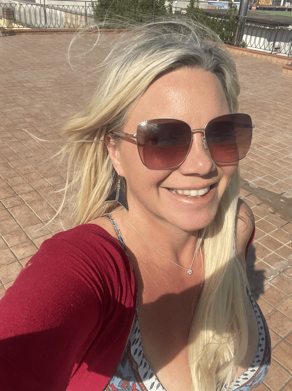 A woman with long blonde hair wearing large sunglasses, a red top, and a patterned dress, smiling outdoors on a sunny day.