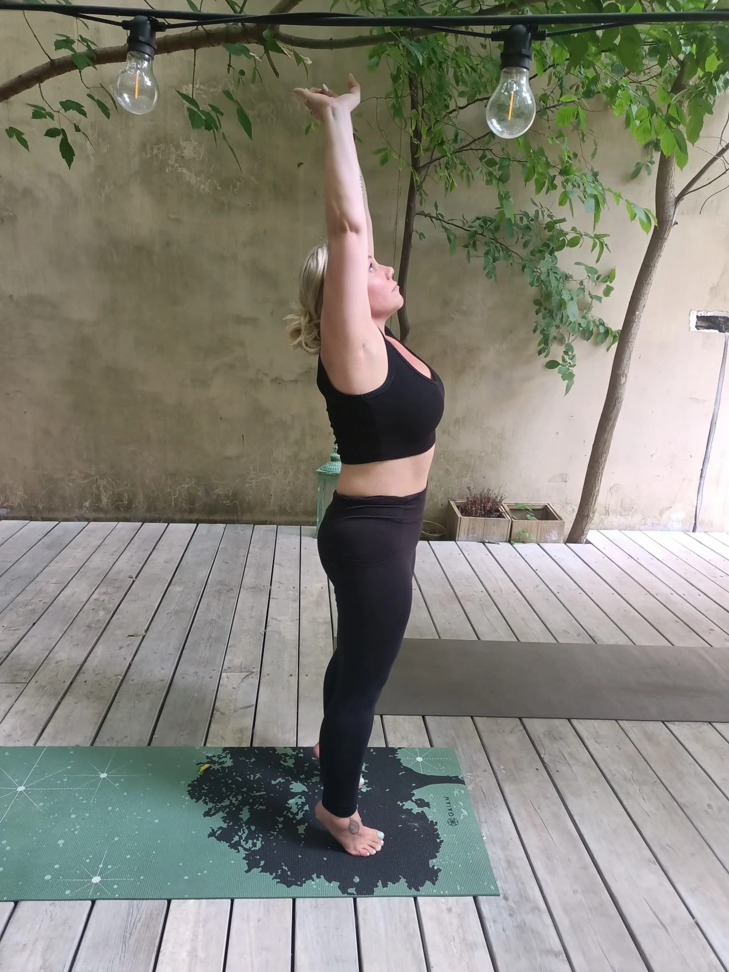 A woman practicing yoga outdoors on a wooden deck, standing in a pose with arms raised above her head, looking upward. She is on a green yoga mat with a tree and string lights overhead.
