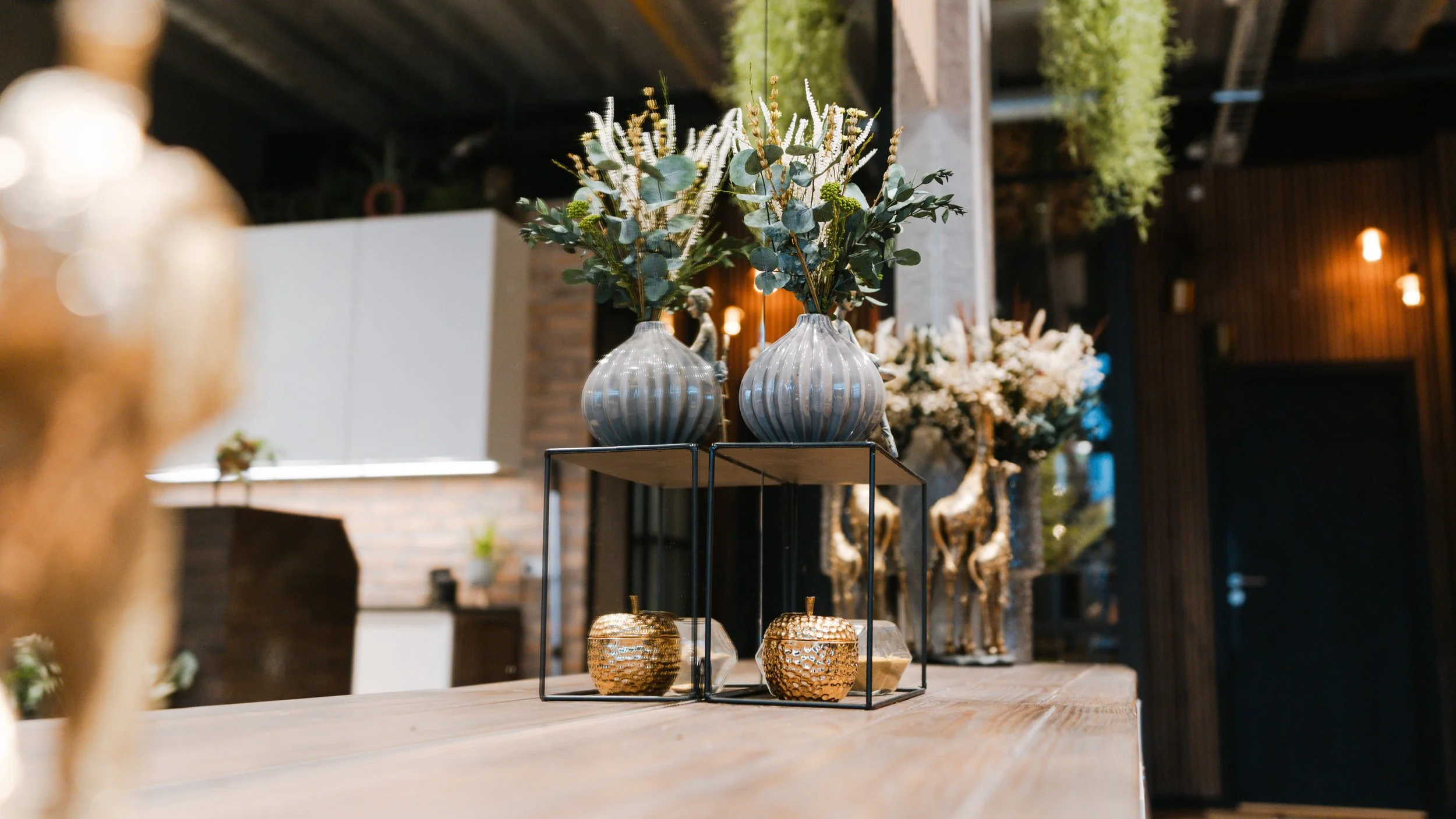 Dekorative vaser med blomster på et træbord i et indendørs miljø med mørkt trævæg og bløde belysning.