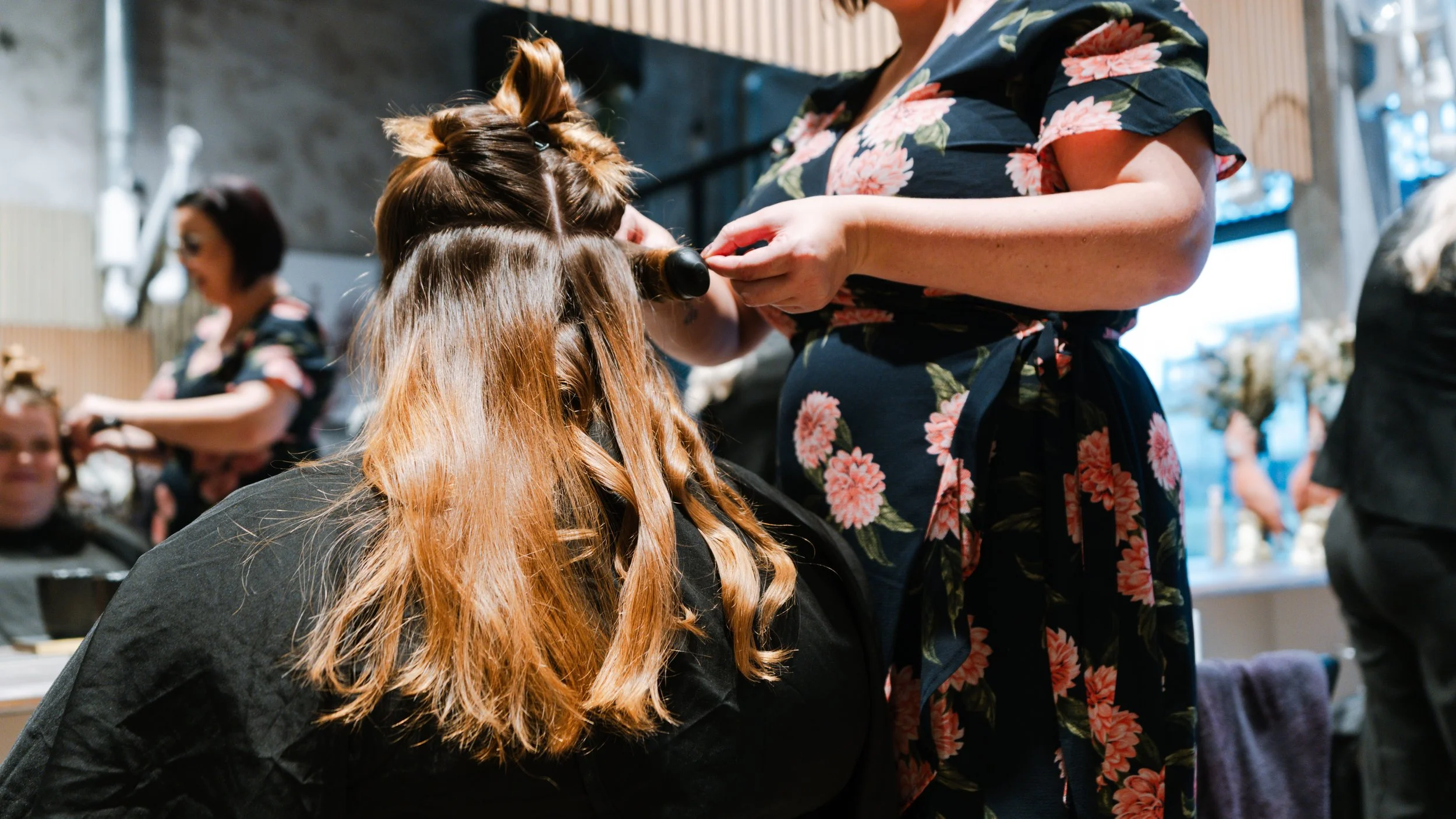 En frisør styler en kvindes lange, lyse hår, mens andre personer også arbejder på kunder i baggrunden.