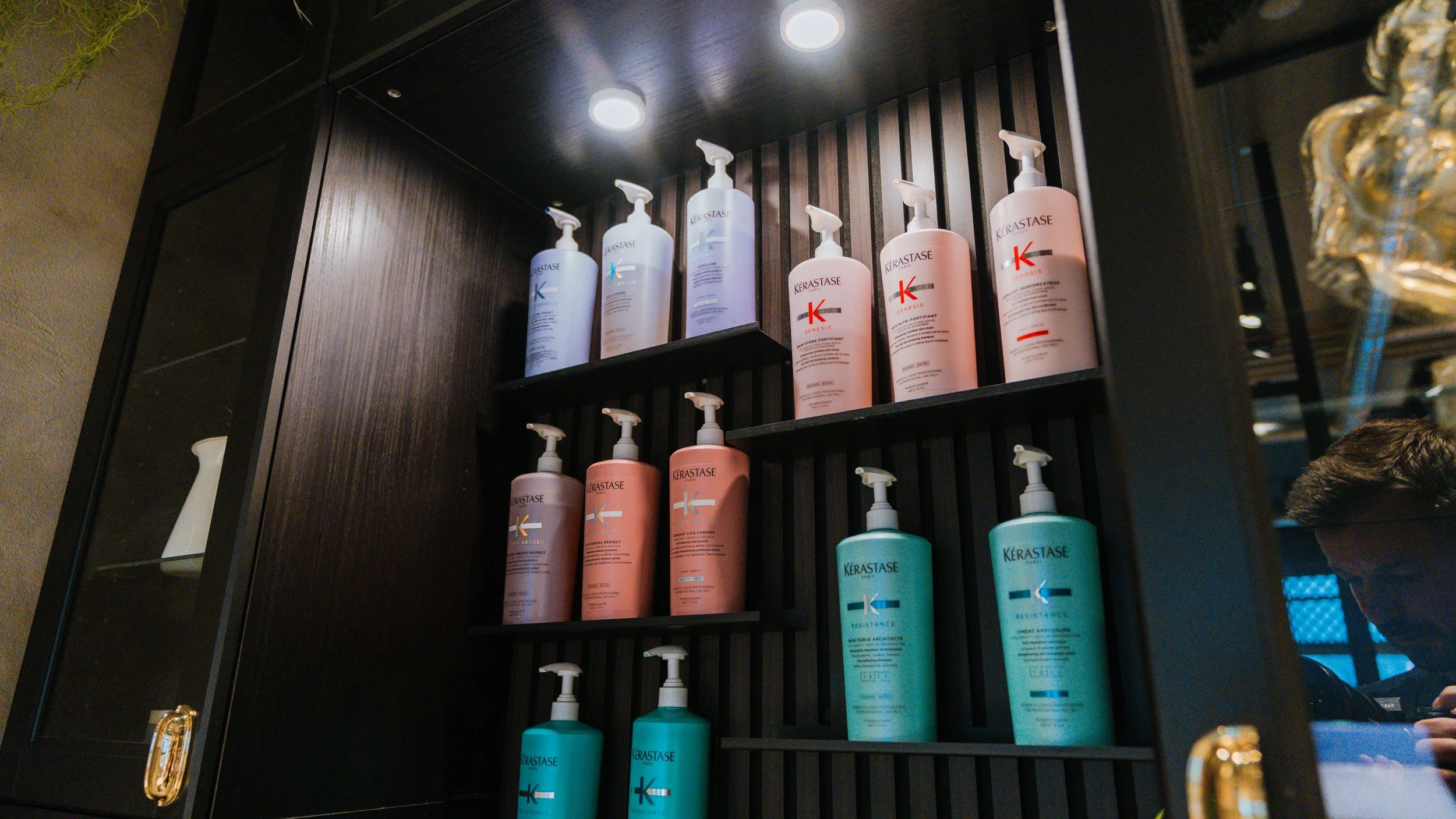 Bøtter med hårplejeprodukter på en sort hylde, inklusive Kérastase Resistance og Genesis mærkede flasker.