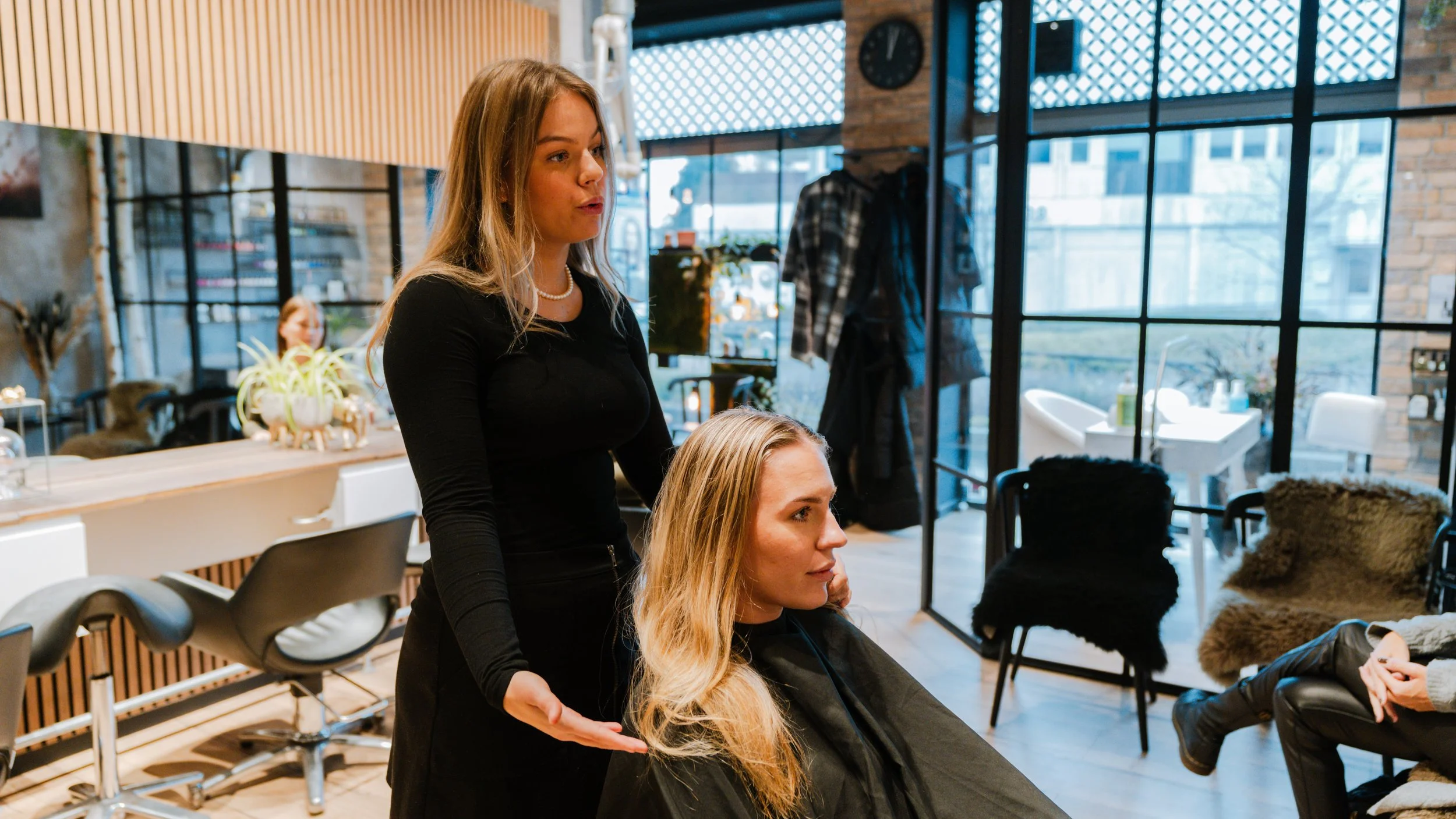 En frisør, en kvinde med blond hår sidder i en frisørstol, mens en anden kvinde med langt blond hår står bag hende i en moderne salon.