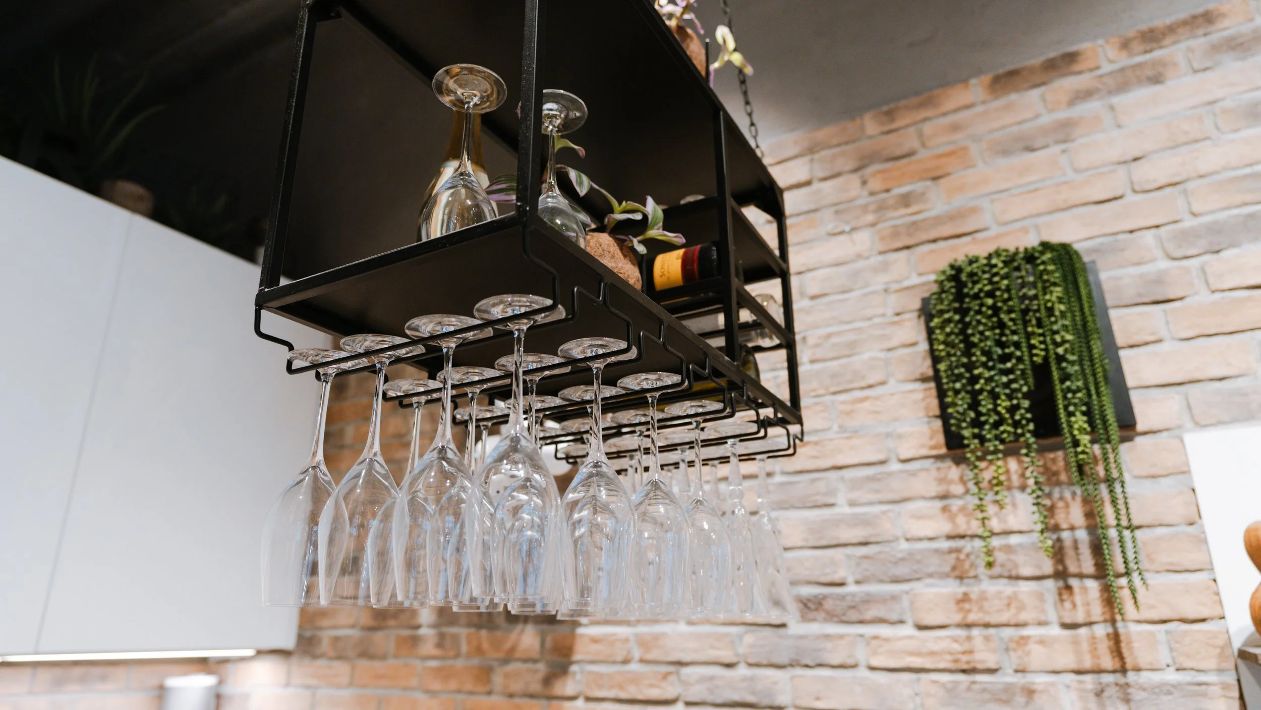 Hængende vinkøleskab med vinglas under, mod en murstensvæg med grøn plante på væggen.