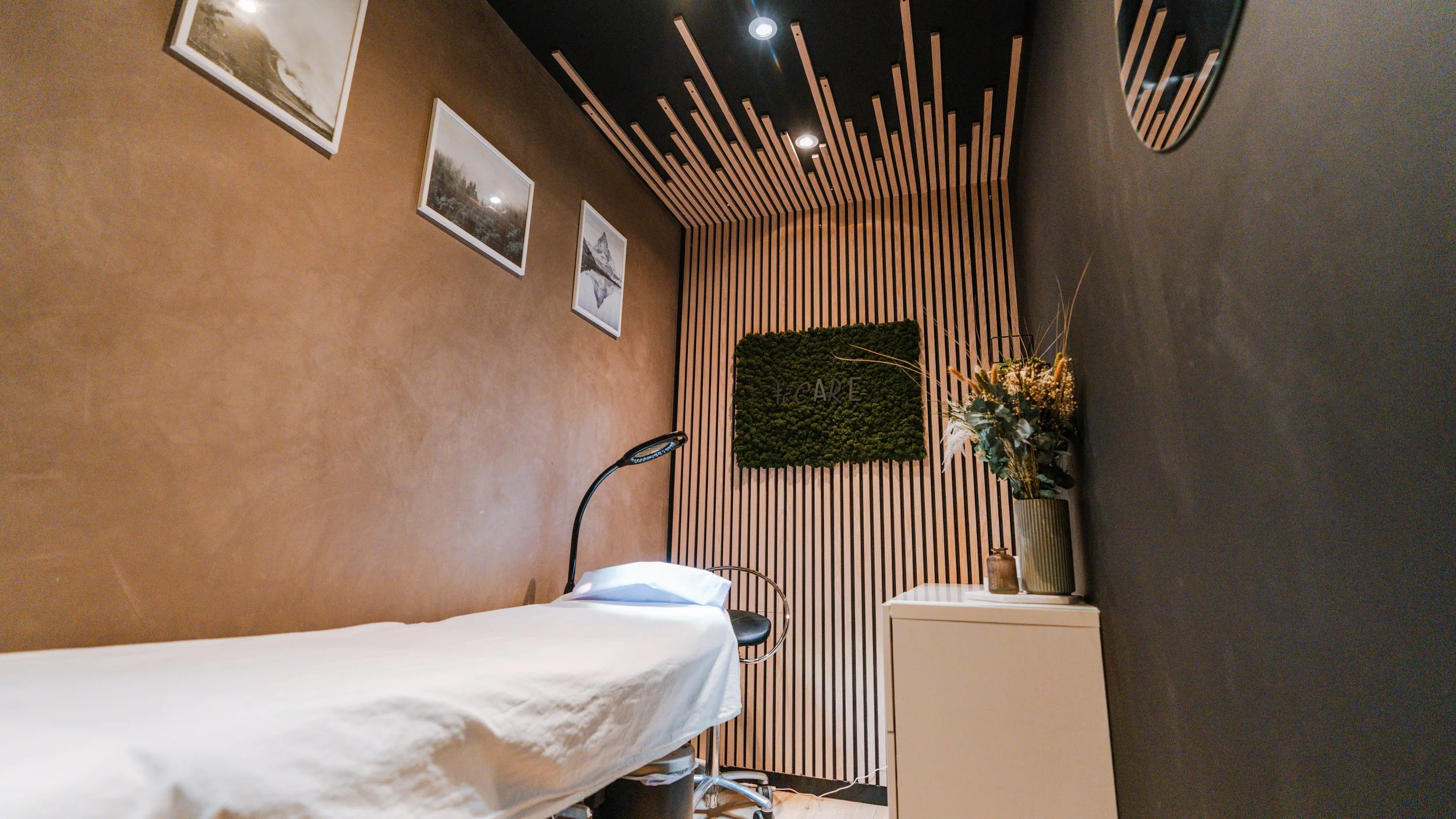 Et behandlingsrum med en enkel seng med hvidt lagen, en sort lænestol, vægge med træpaneler og billeder, og en stor vase med blomster.