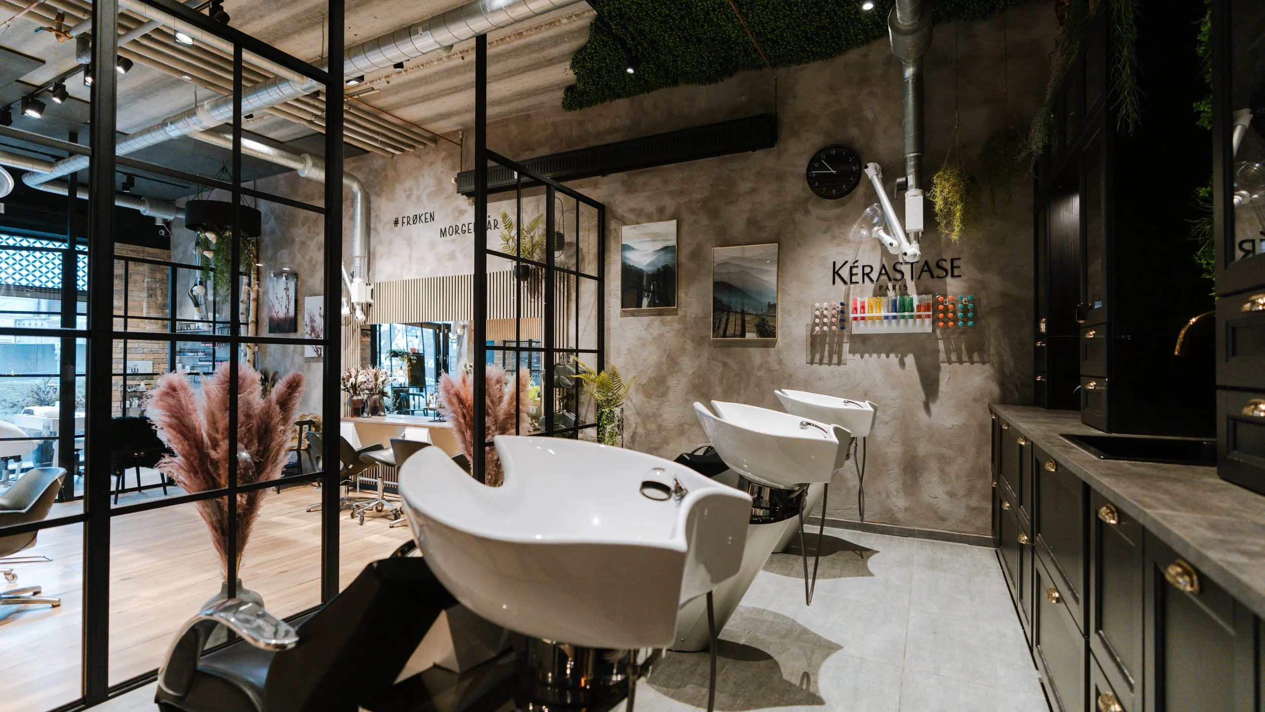 Indretning af en frisørsalon med hårvaskebægre, spejle, grønne planter, og kunstværker på væggene.