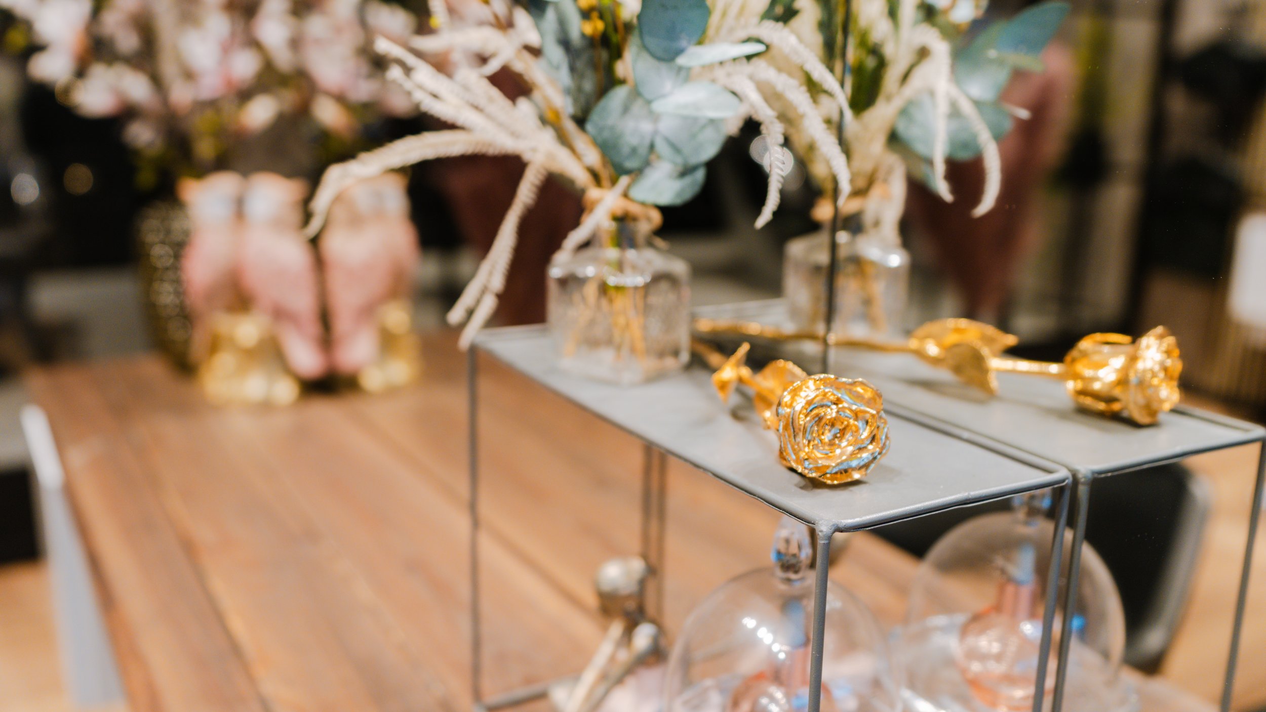 Dekorative blomster i glasvaser på en metalhylde med gyldne roser, foran en træbord.