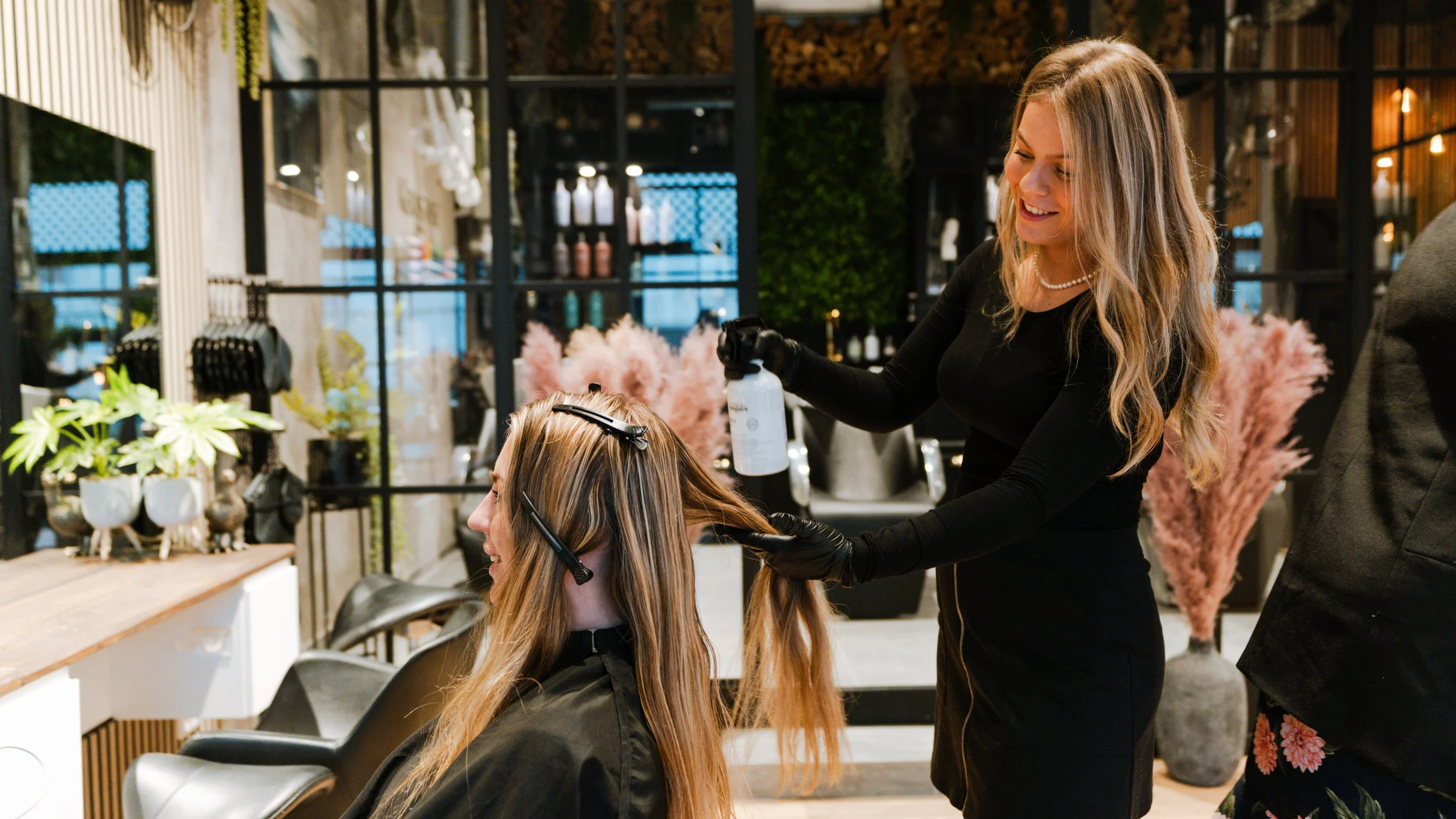 En frisør laver hårfarvning på en kunde i en moderne frisørsalon. Kunder sidder i sorte frisørstole med beskyttende kapper, mens frisøren bruger spray og handsker.
