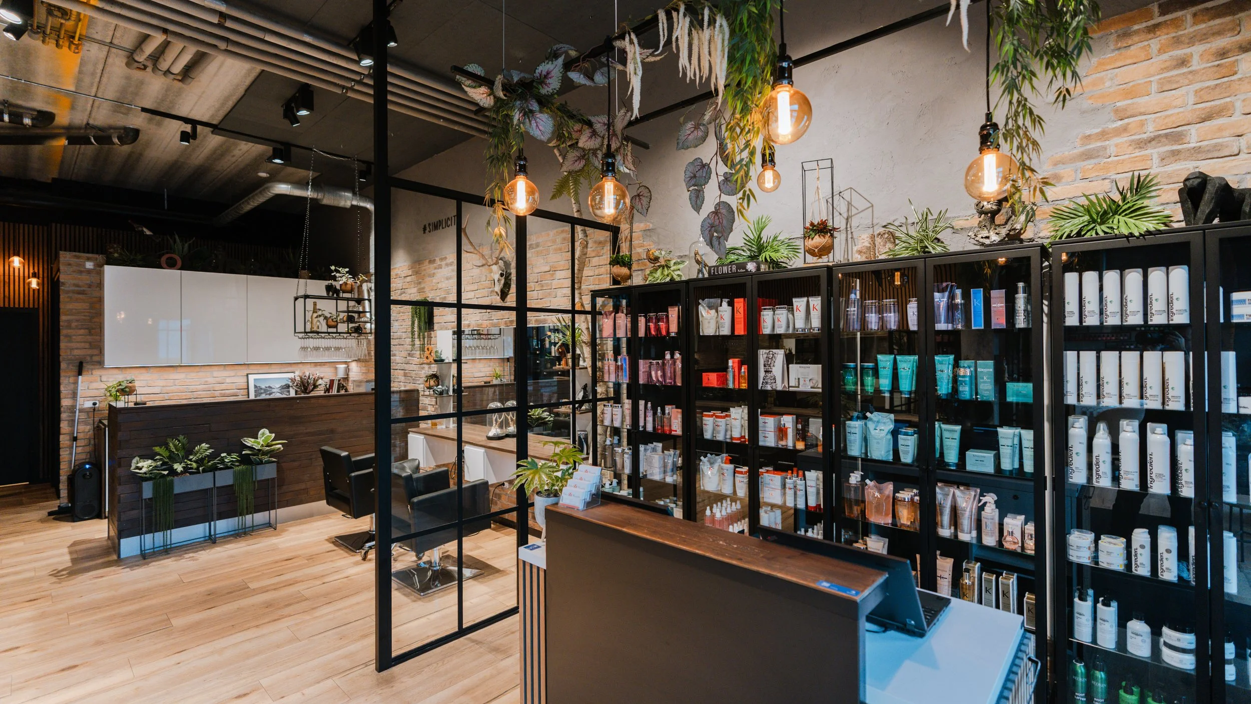 Indretning af en frisørsalon eller skønhedssalon med hylder fyldt med produkter, planteudsmykning, pærer og en receptionist-område.