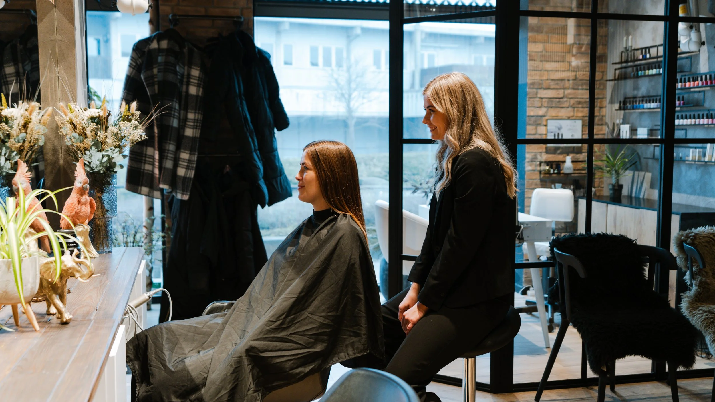 En kvinde sidder i en frisørstol med en kappe, mens en anden kvinde står foran hende og smiler, i en moderne salon med glasvægge og hylder med neglelak.