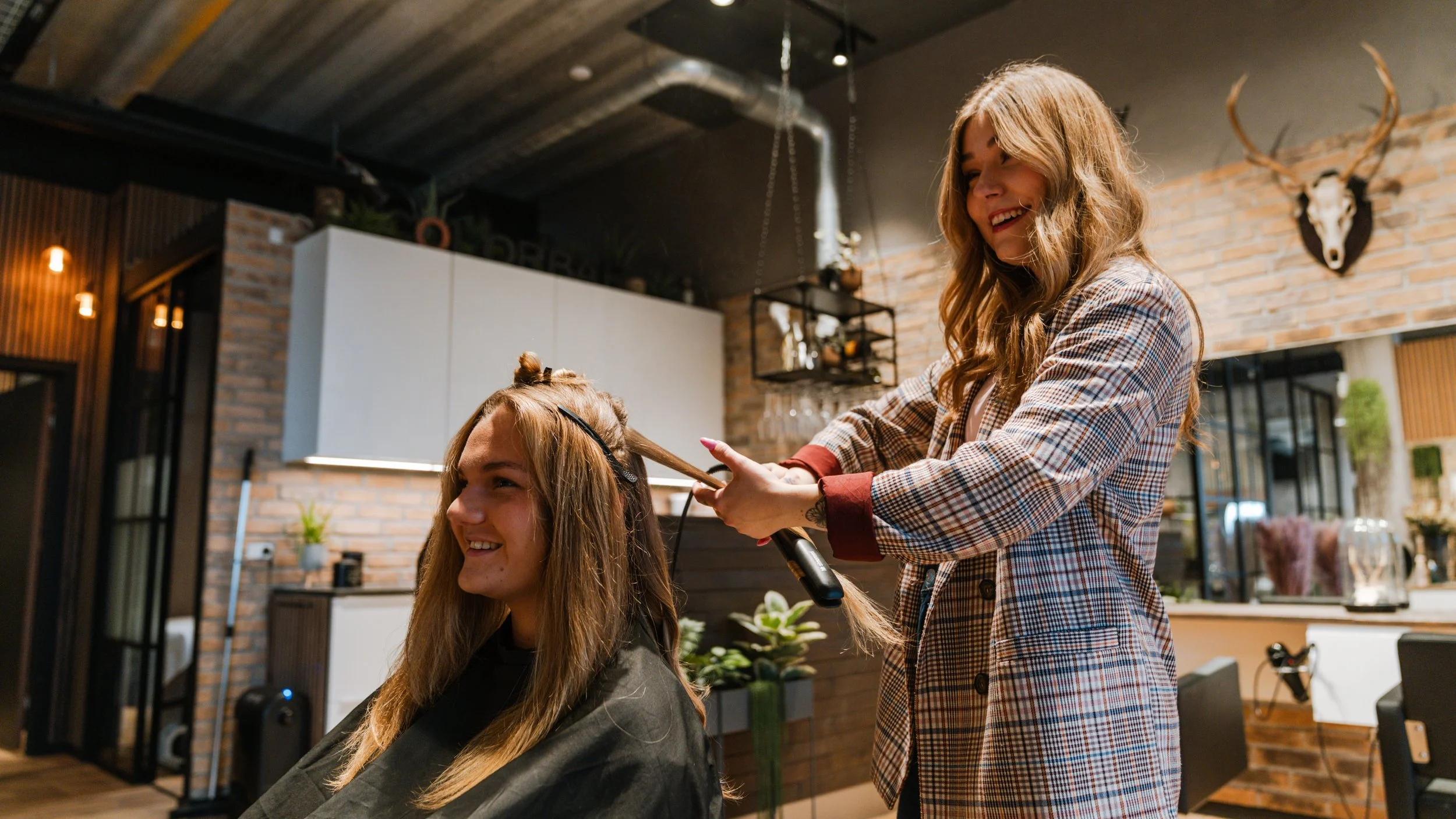 En frisør, der klipper en kvindes hår i en trendy salon med moderne indretning, brændte murstensvægge og dekorative dyreskaller på væggen.