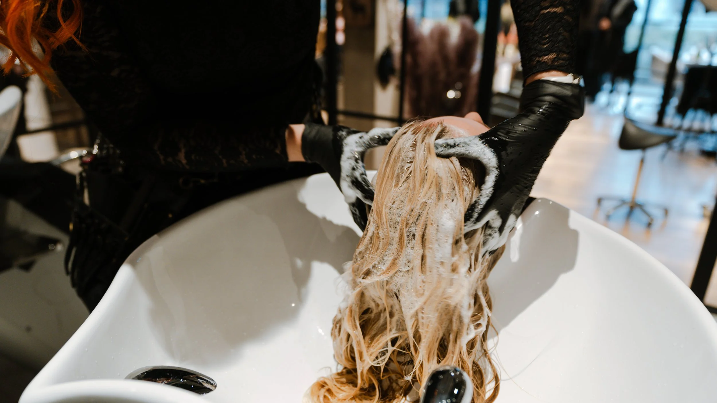 En frisør vasker en kundes blonde hår i en frisørstolvask med sort handsker.
