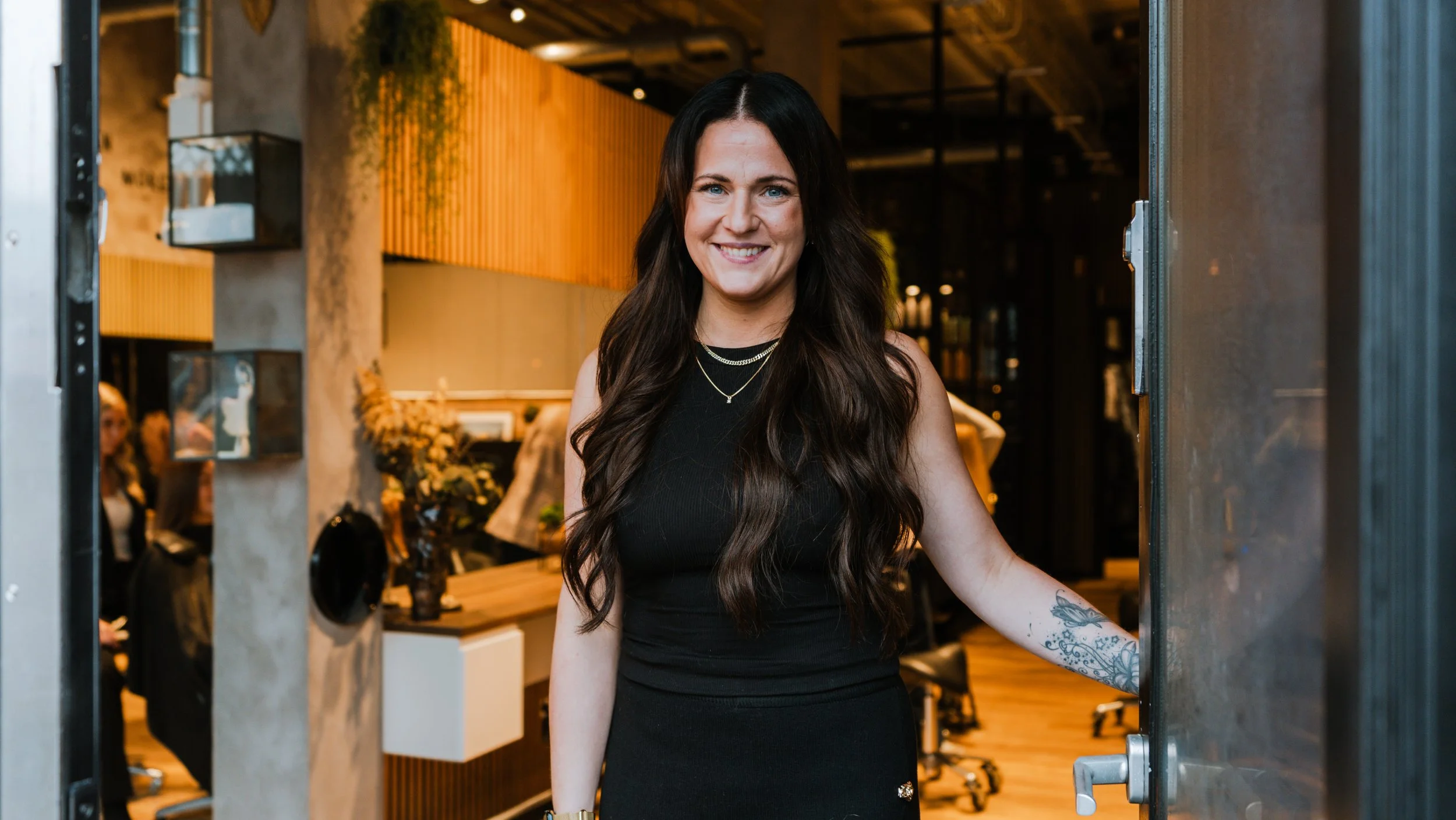 En smilende kvinde med langt, brunt hår står i en dørramme indendørs, med en moderne indretning i baggrunden.