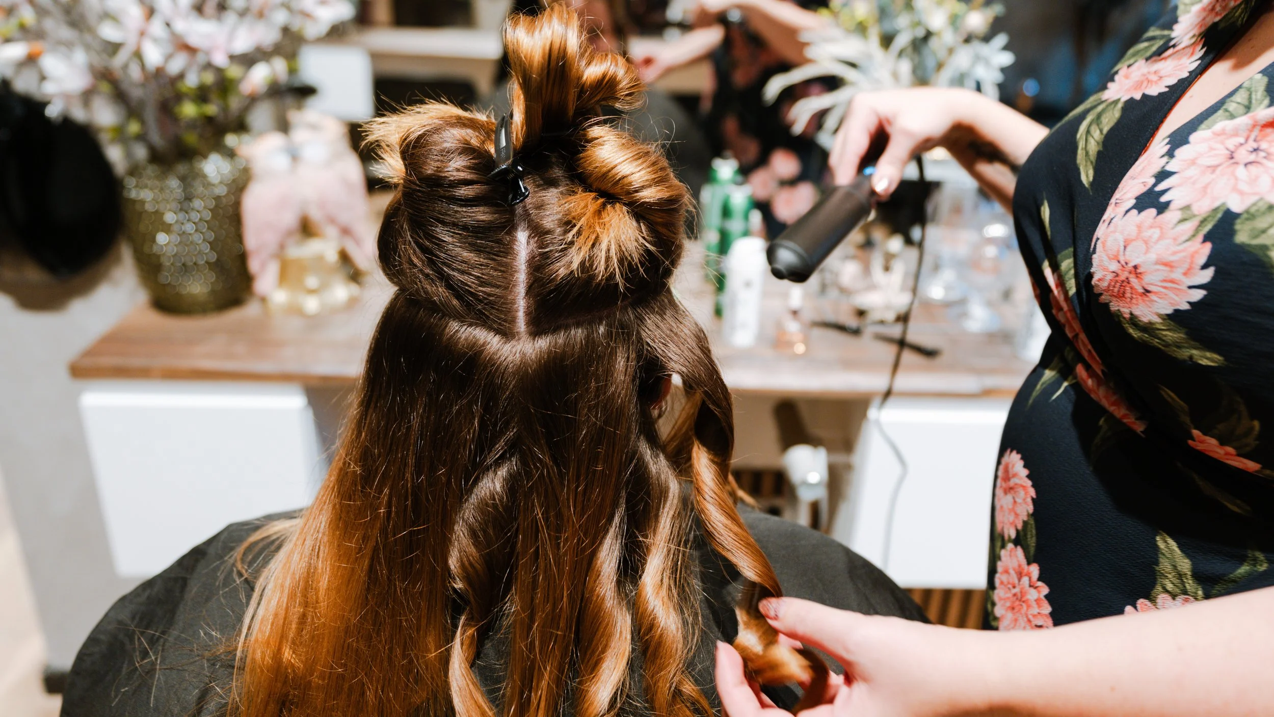 En frisør, der bliver klippet og stylet af en person, mens en anden hjælper, i en frisørsalon med blomsterdekorationer på væggen og forskellige hårprodukter på bordet.