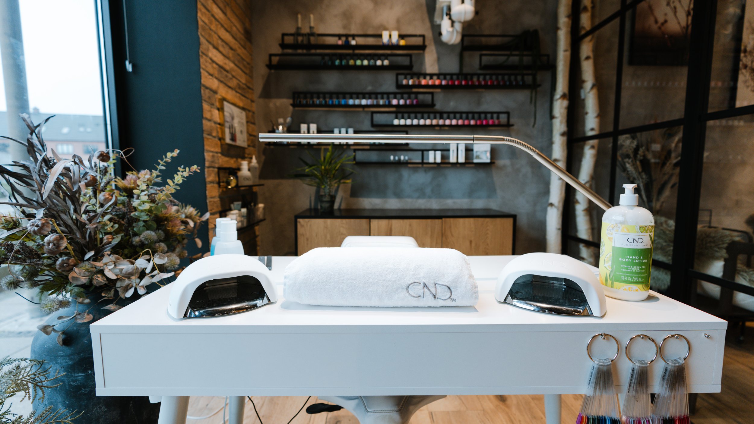 Et manicurebord med UV-lamper, håndklæde med logo, hånd- og kropslotion, og negleværktøjer, i en salon med moderne indretning.