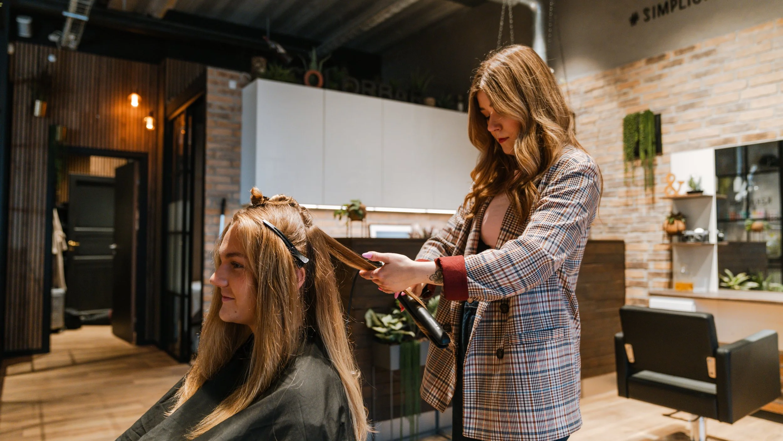 En frisør klipper en kvinde i en frisørsalon med industrielle design, træ- og murstensvægge.