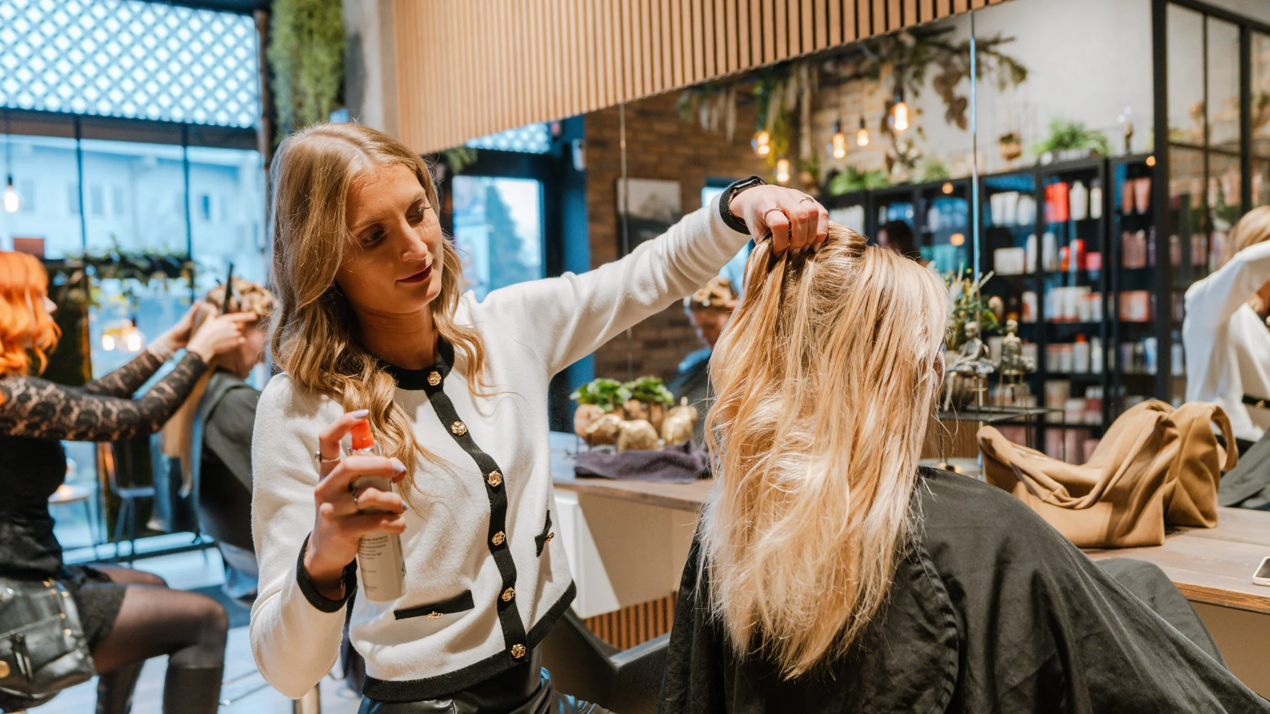 En frisør, der holder en hårspray i hånden, arbejder på en kvinde med blondt hår i en salon.