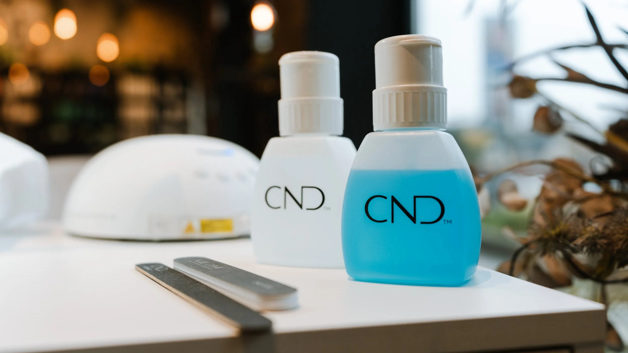 To flasker med blå og hvide labels med mærket 'CND' placeret på en hvid bordplade. I baggrunden er der et integreret UV-lysapparat og noget planteværk.