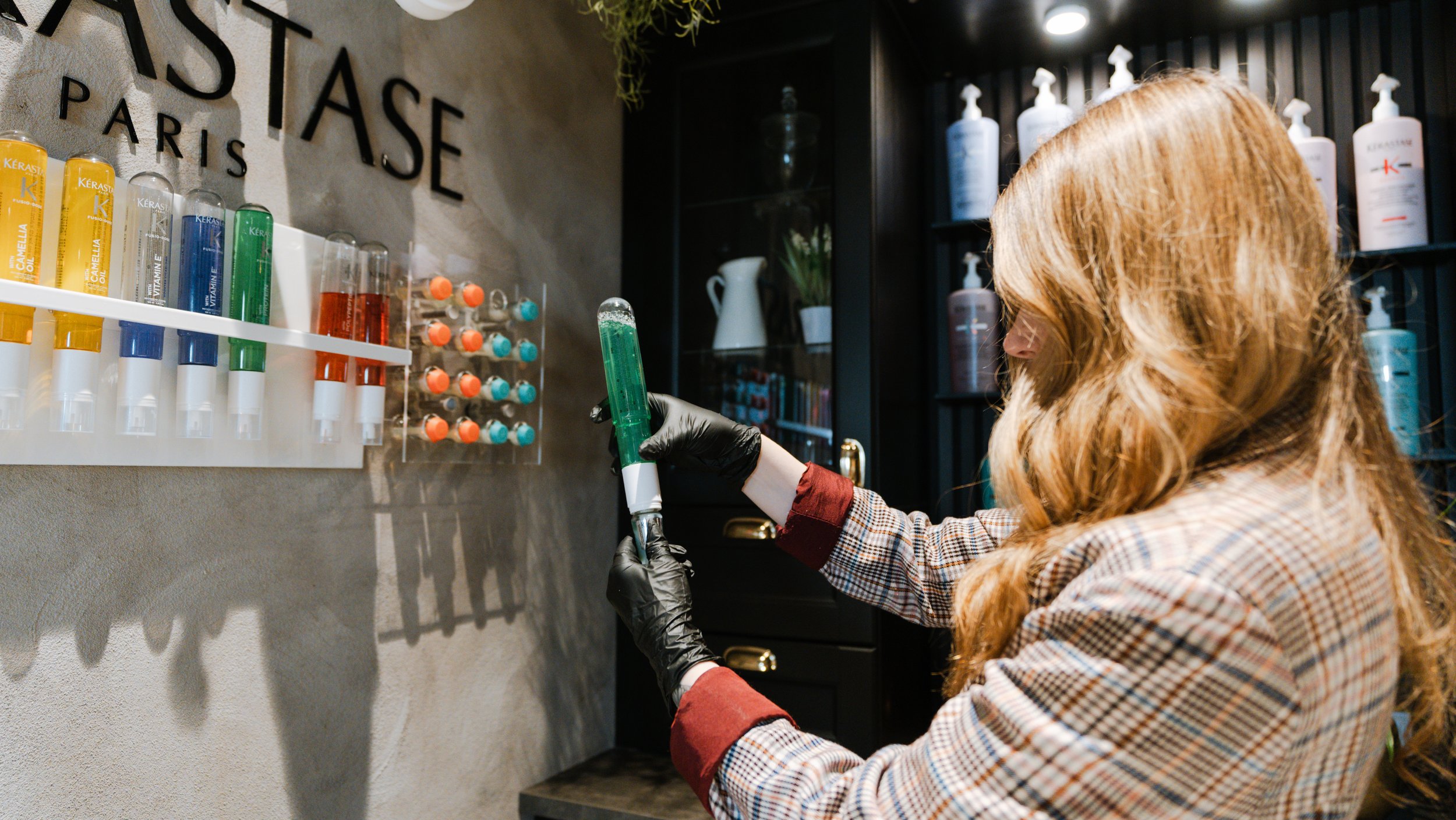 Kvindelig person med rødt hår, der bruger beskyttelseshandsker og håndterer en stor prøve af grøn gel i et laboratorium eller en skønhedssalon, med hylder med shampoo og hårplejeprodukter i baggrunden.