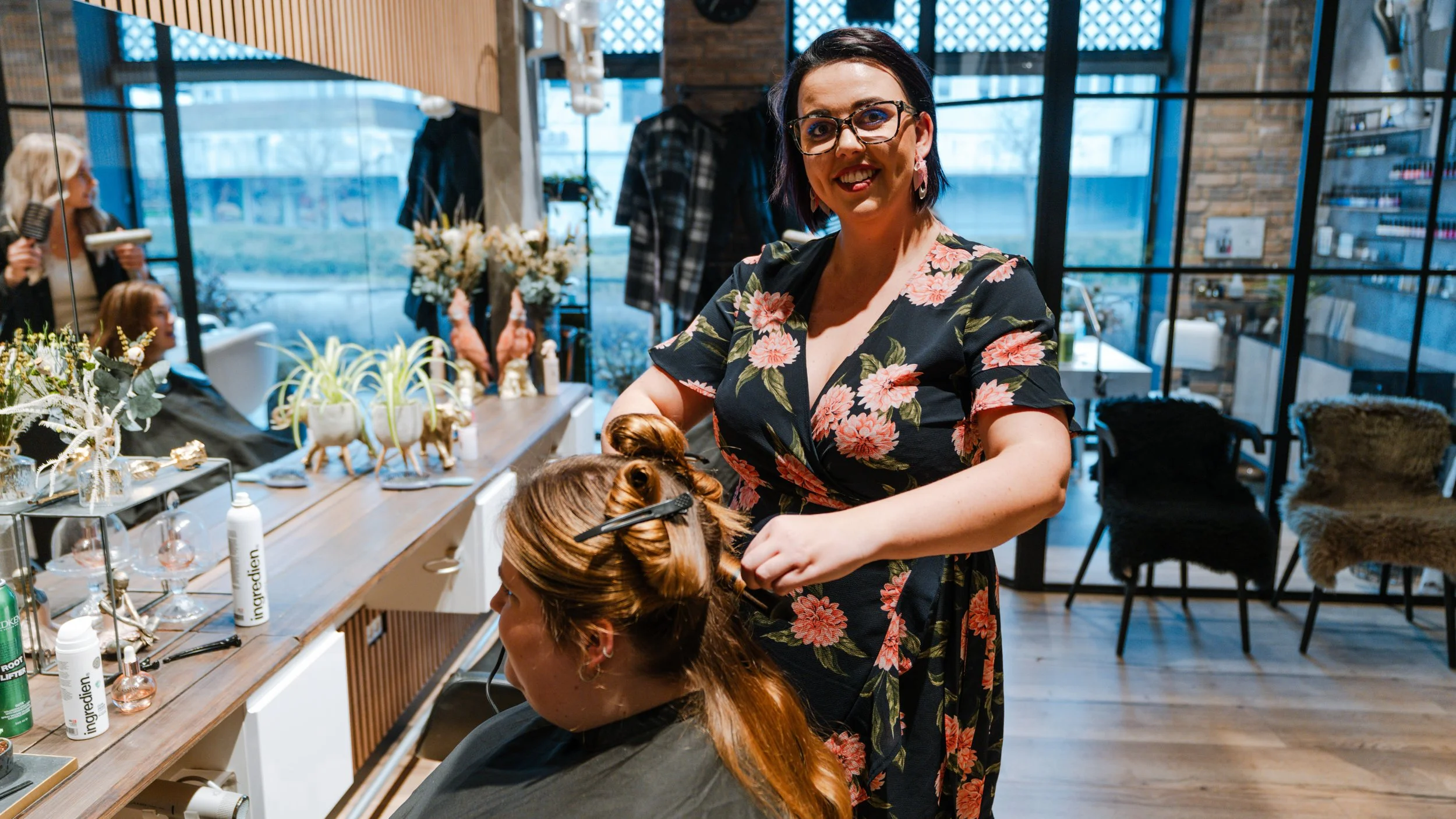 En kvinde med briller og blomsterprint kjole giver en klipning til en anden kvinde, der sidder i en stol i en frisørsalon.