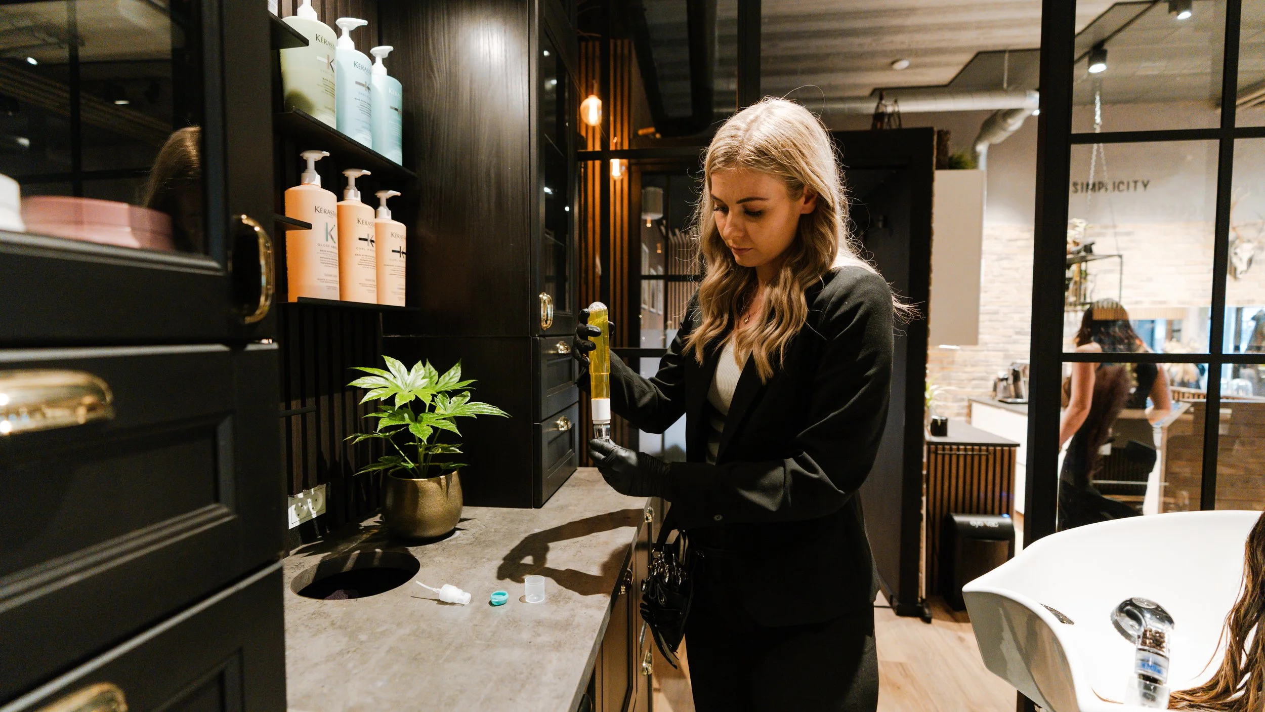 En kvinde iført sort jakke og handsker, der håndterer en tube med hårfarve i en frisørsalon med hylder med shampoo og en grøn plante.