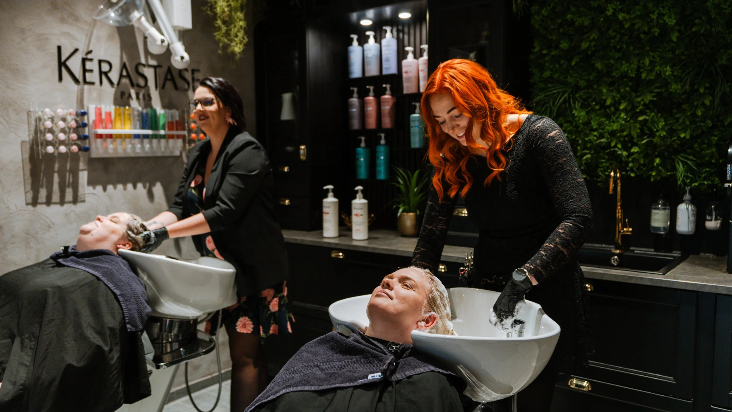 To frisører vasker kunders hår i en salon med mørk indretning og grønne planter.