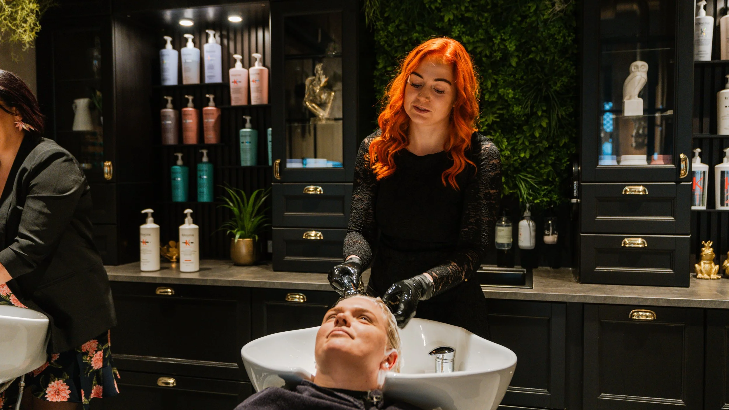 En frisør klipper en mands hår i en salon med sort indretning og grønne plantevægge.