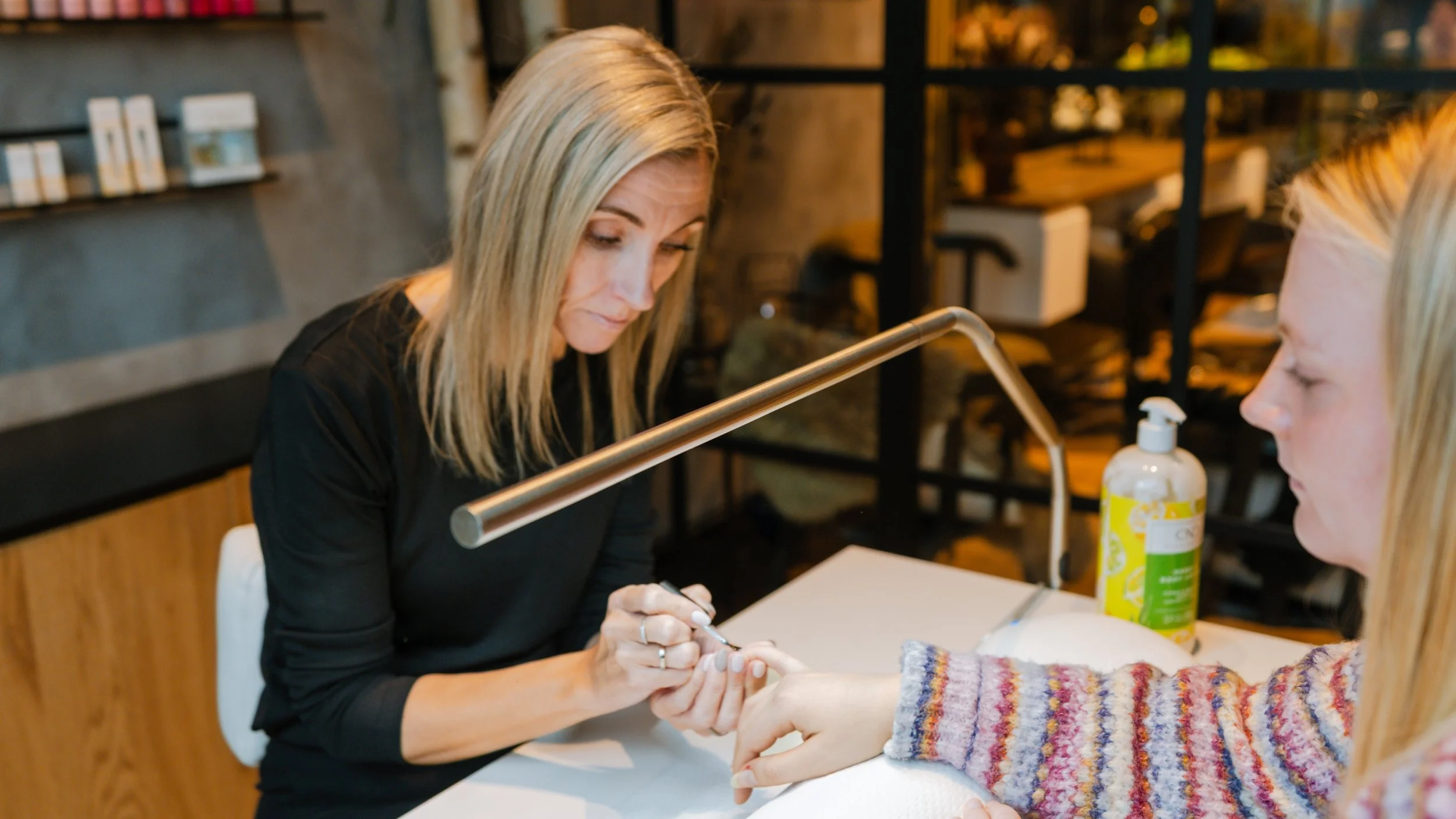 En kvinde får manicure af en anden kvinde i en salon med moderne indretning og hylder med produkter i baggrunden.