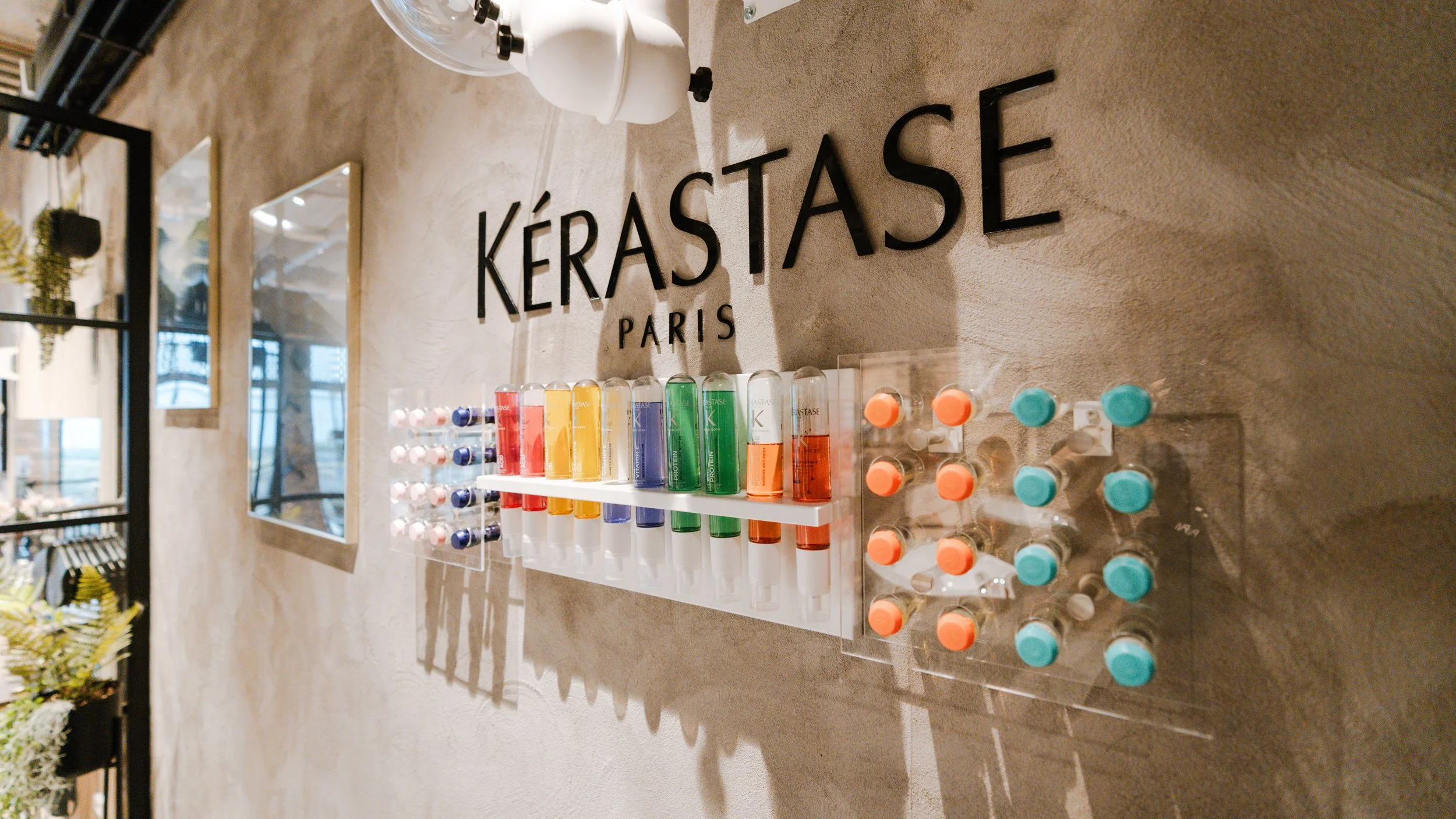 interiørfoto af en væg med Kerastase Paris logo og forskellige farvede hårprodukter i rør, samt små flasker med orange og blå tops