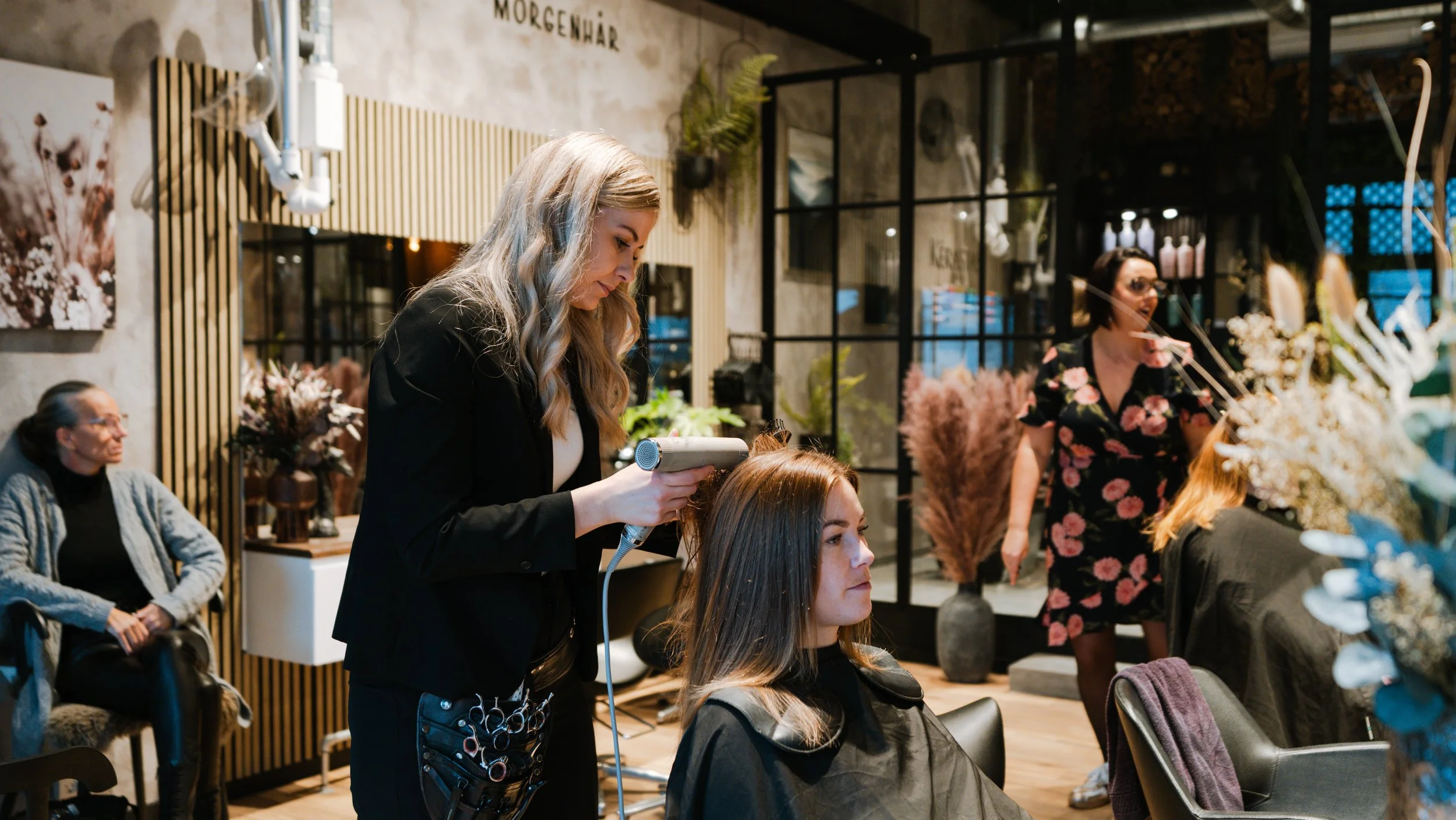 En frisørsalon med flere personer, hvor en frisør klipper en kvindes hår, mens andre sidder og venter. Salonindretning med blomsterdekorationer og store vinduer.