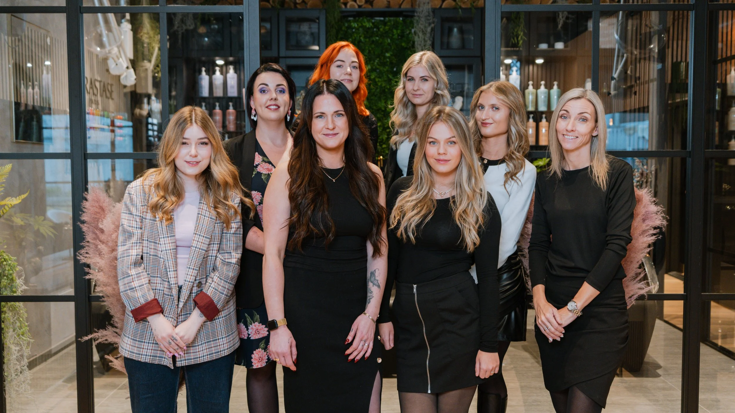En gruppe på ni kvinder står sammen indendørs foran en glasvæg med hylder fyldt med produkter, smilende og posere for kameraet.