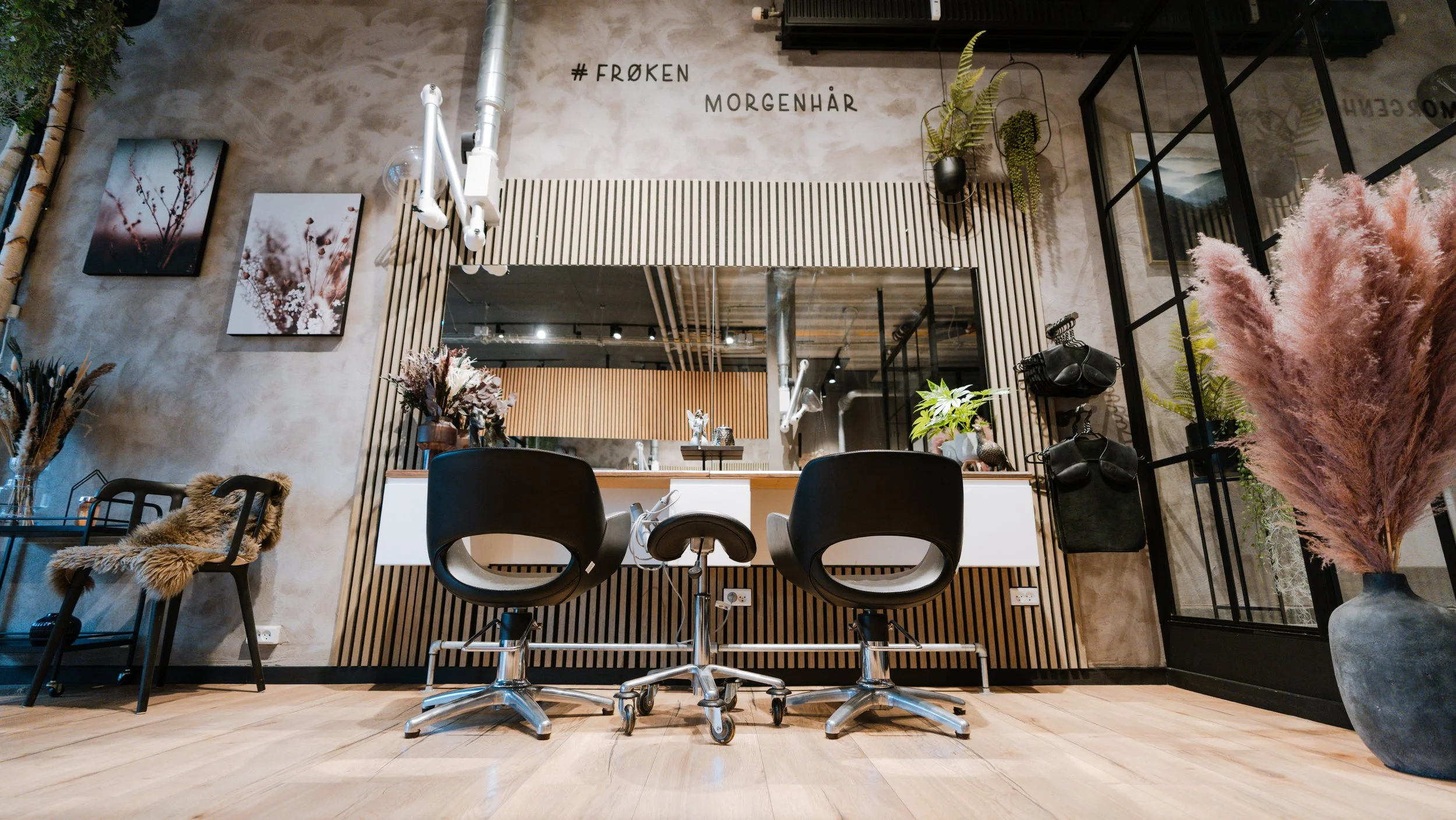 Indretning af en moderne, stilfuld frisørsalon med to sorte kontorstole foran en spejlskærm, blomsterdekorationer, fotografier på væggen, og tekst på væggen der siger '#FRØKEN MORGENHÅR'.