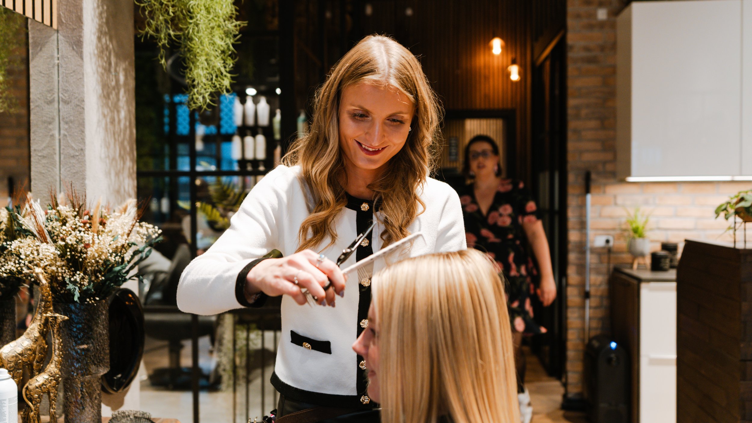 En frisør, der klipper en kundes hår i en salon, med blomsterdekorationer på bordet og en kvinde i baggrunden.