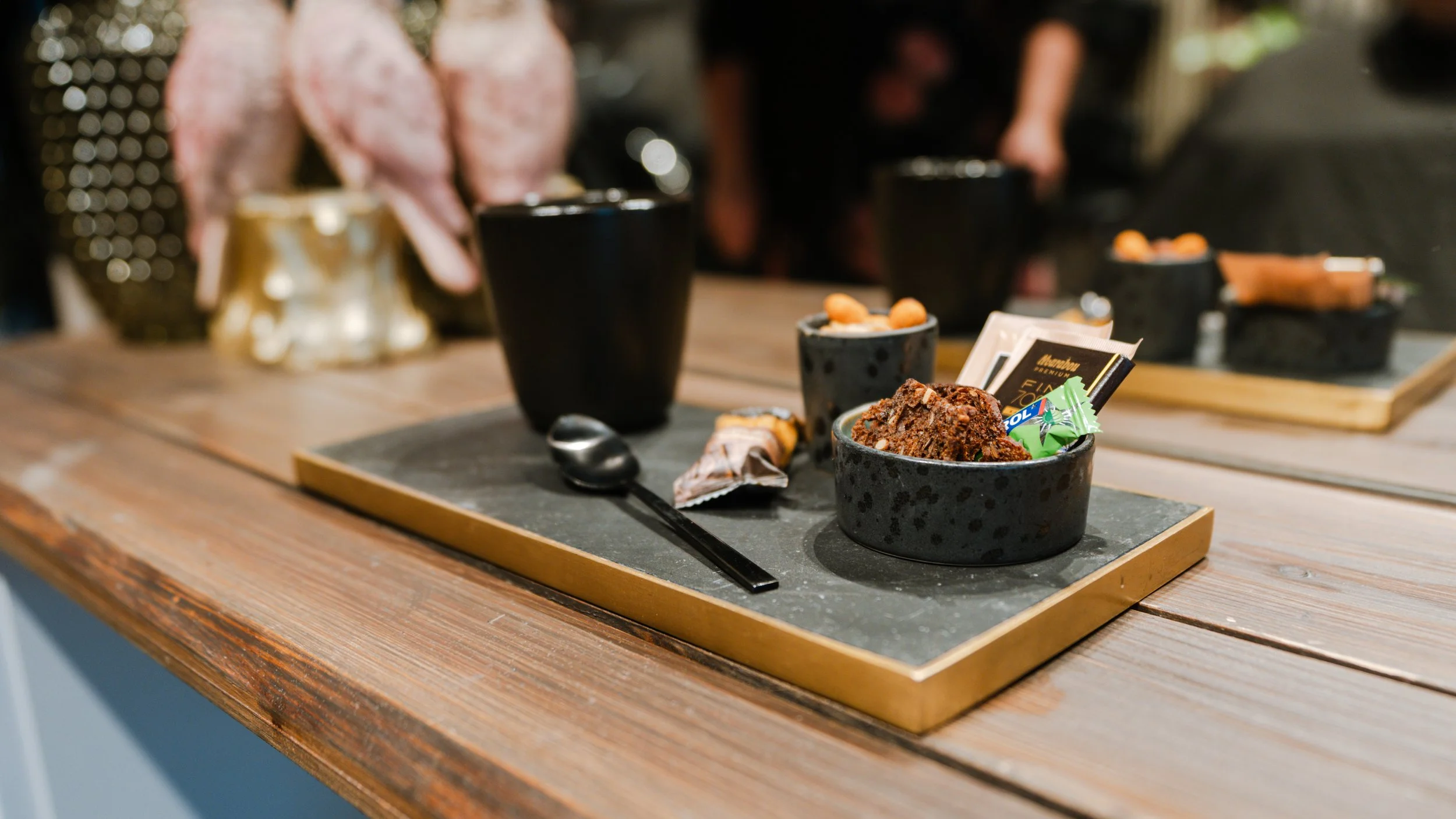 Billedet viser en tallerken med chokolade brownie, små pakker med slik, en sort kop med en dessert, to sorte kopper med snacks og en ske på en træbord.