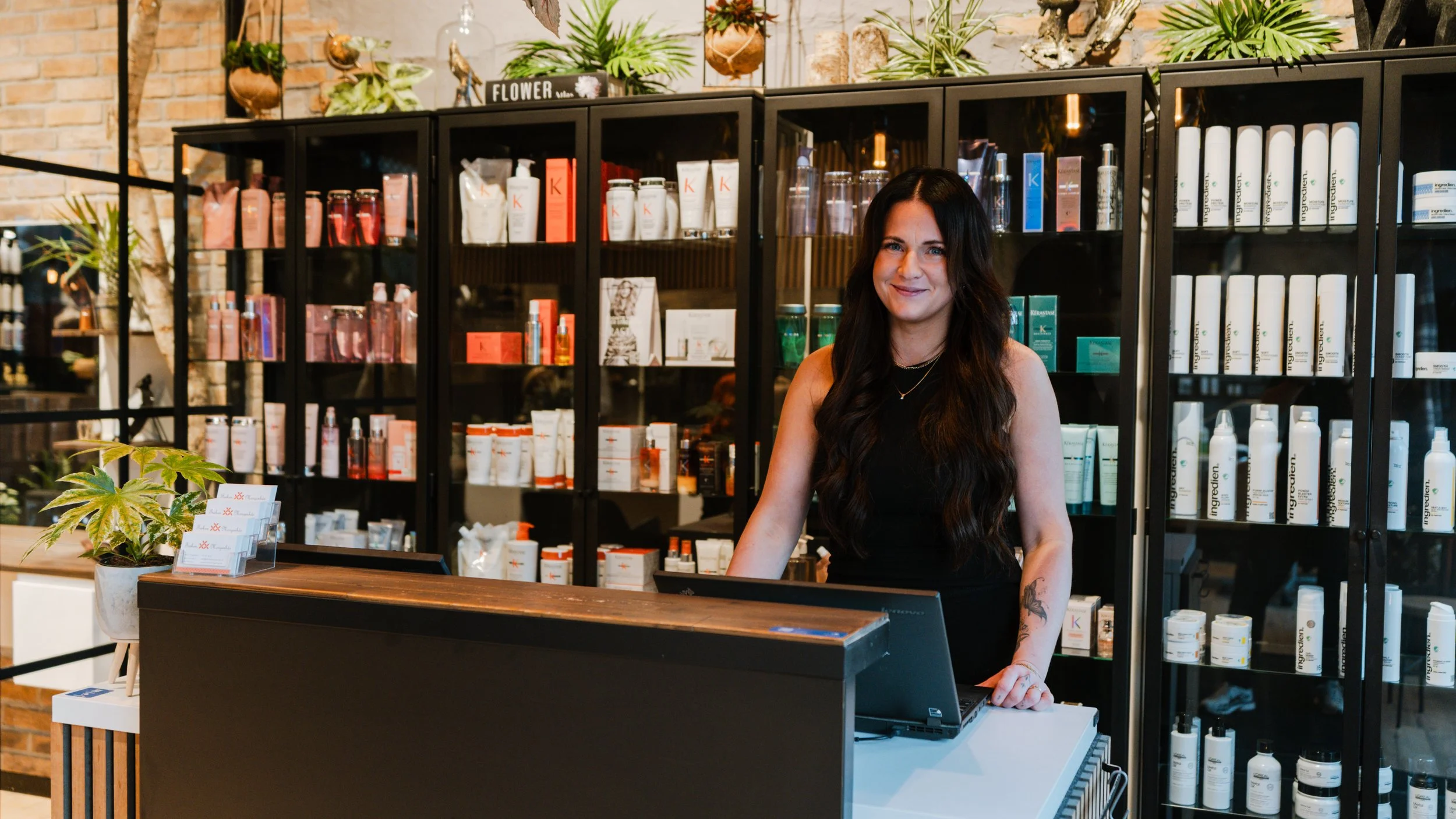 En kvinde med mørkt, krøllet hår står bag en kasse i en frisørsalon, omgivet af hylder med frisør- og skønhedsprodukter.