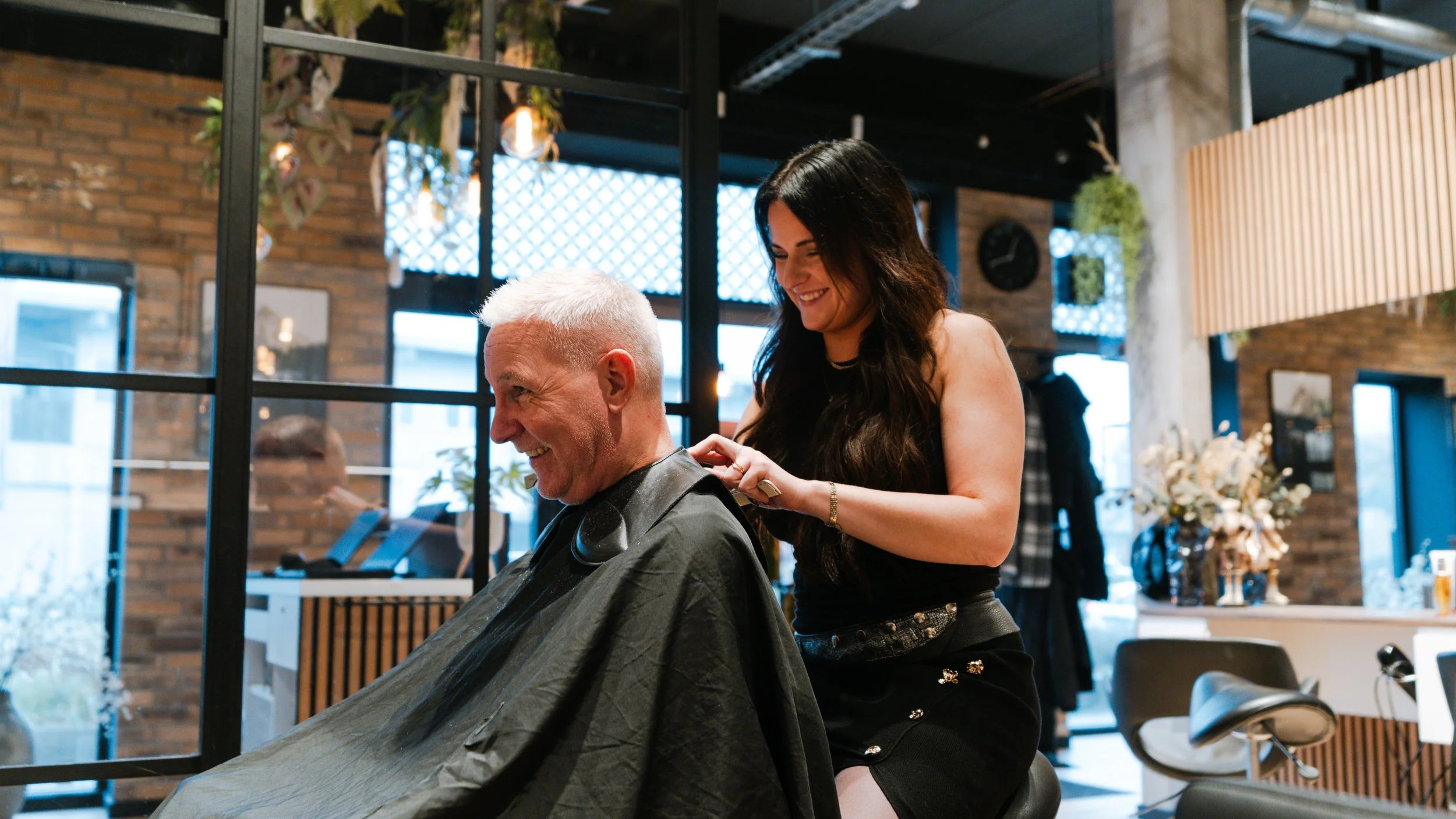 En mand sidder i en frisørstol med klipning, mens en kvindelig frisør står bag ham og klipper ham i nakken, i en moderne frisørsalon.