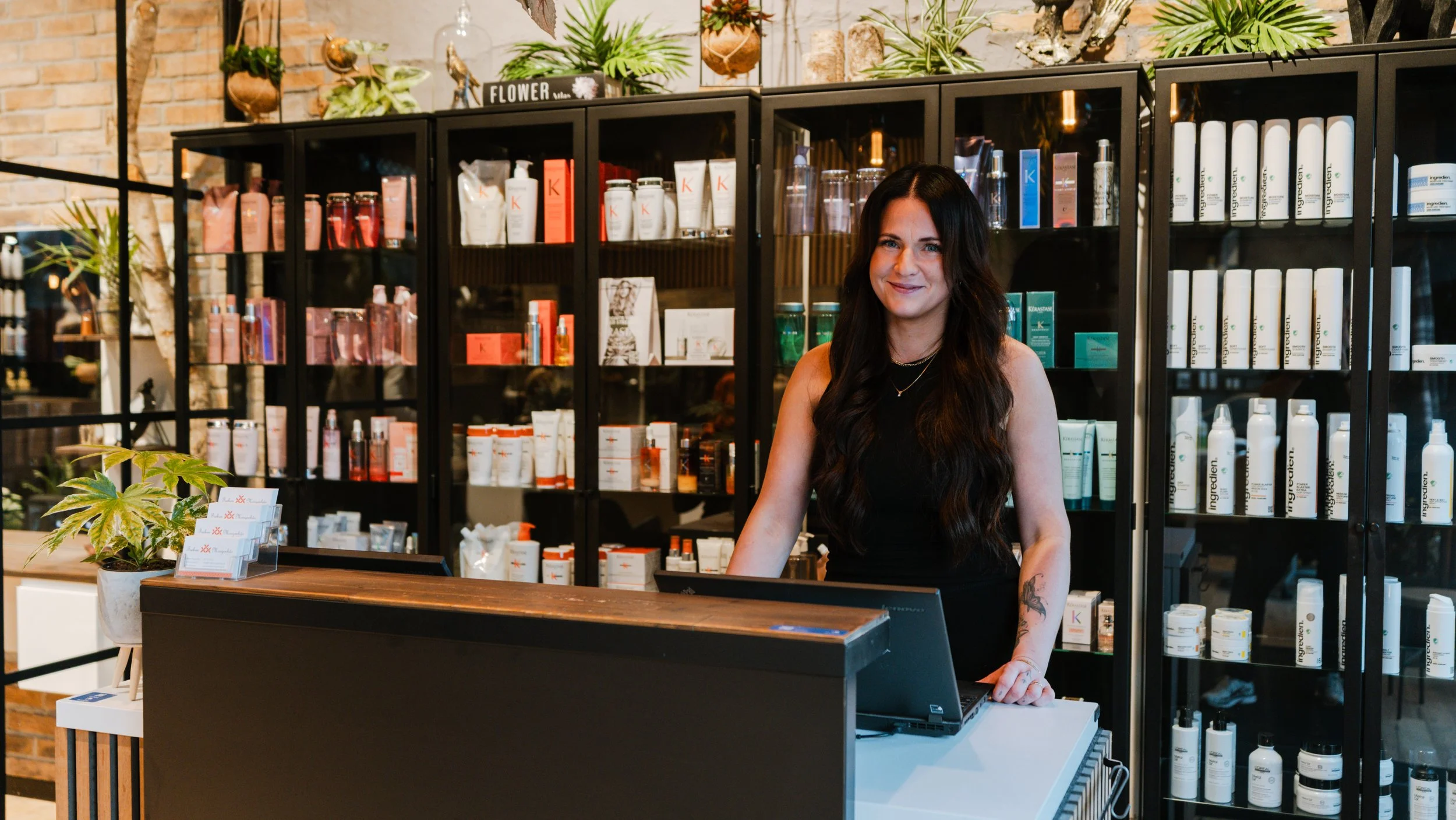 Kvindelig indehaver bag skranken i frisørsalon med sorte hylder bagved, fyldt med frisør- og skønhedsprodukter.