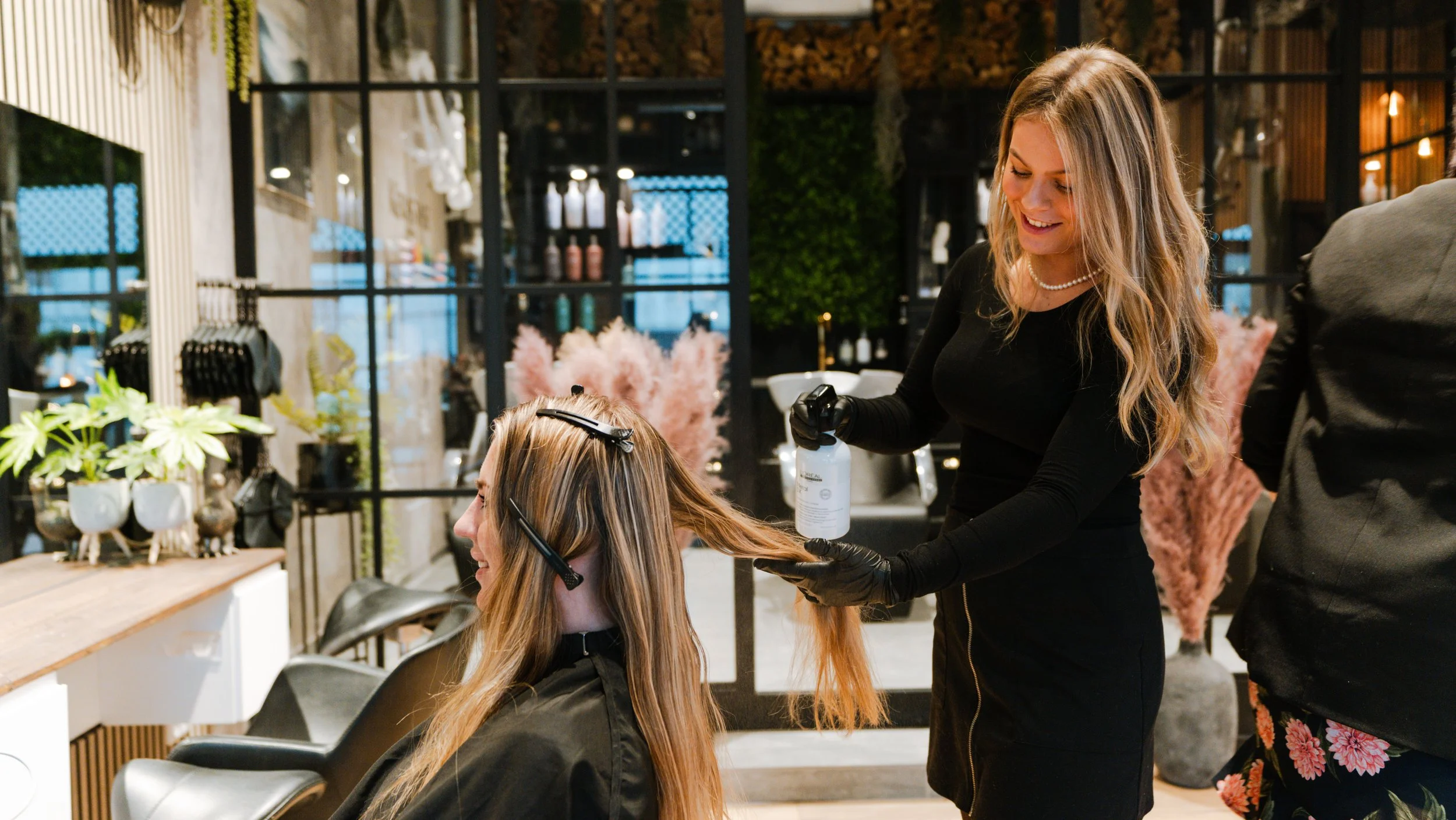 En frisør, der sprøjter hår med hårprodukter, er ved at style en kvindelig kunde i en moderne frisørsalon med indretning af planter og dekorative elementer.