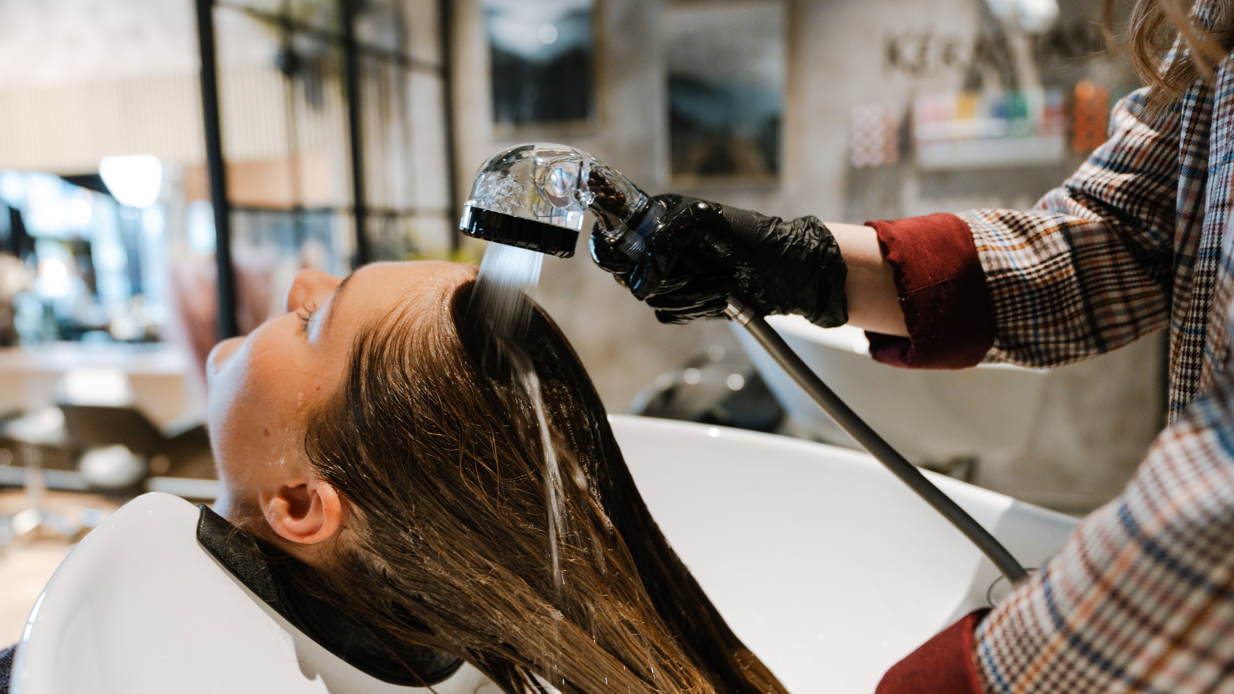 En kvinde sidder med hovedet tilbage i en vask, mens en person vander hendes lange brune hår i en frisørsalon.