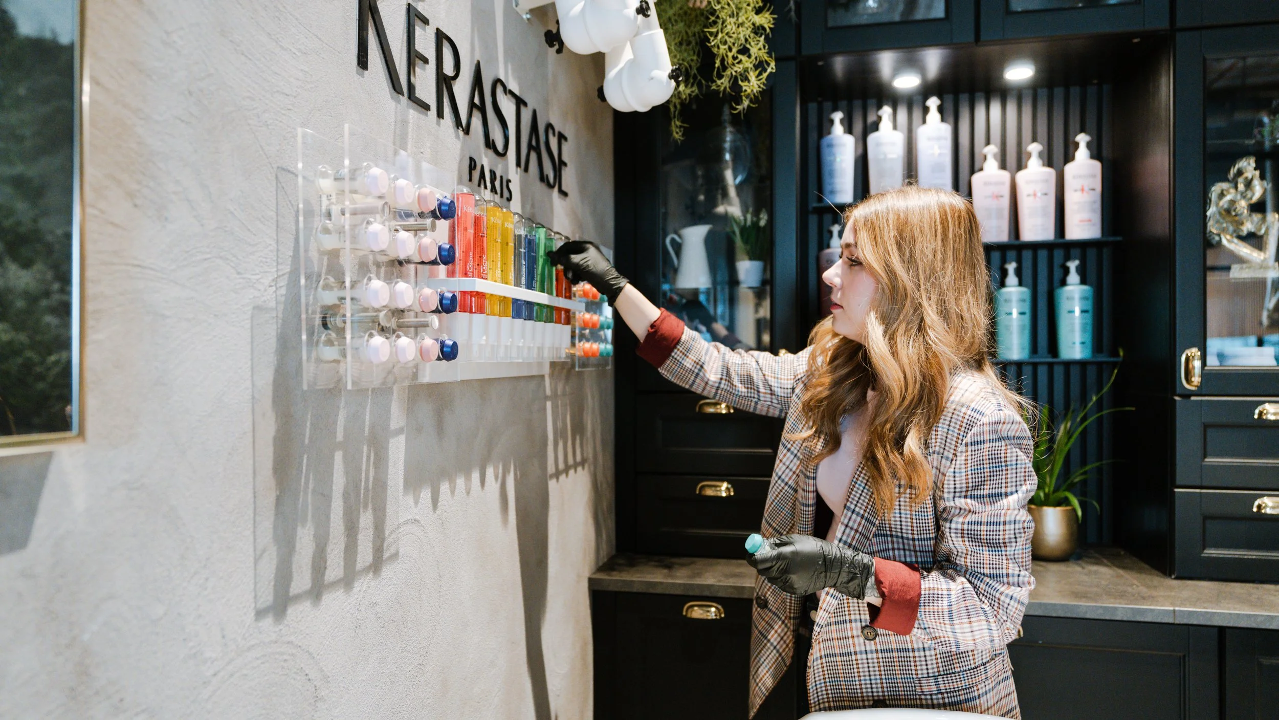 Kvinde med brunt, bølget hår, iført sort handske, vælger neglelak fra farvehjul i skønhedssalon.