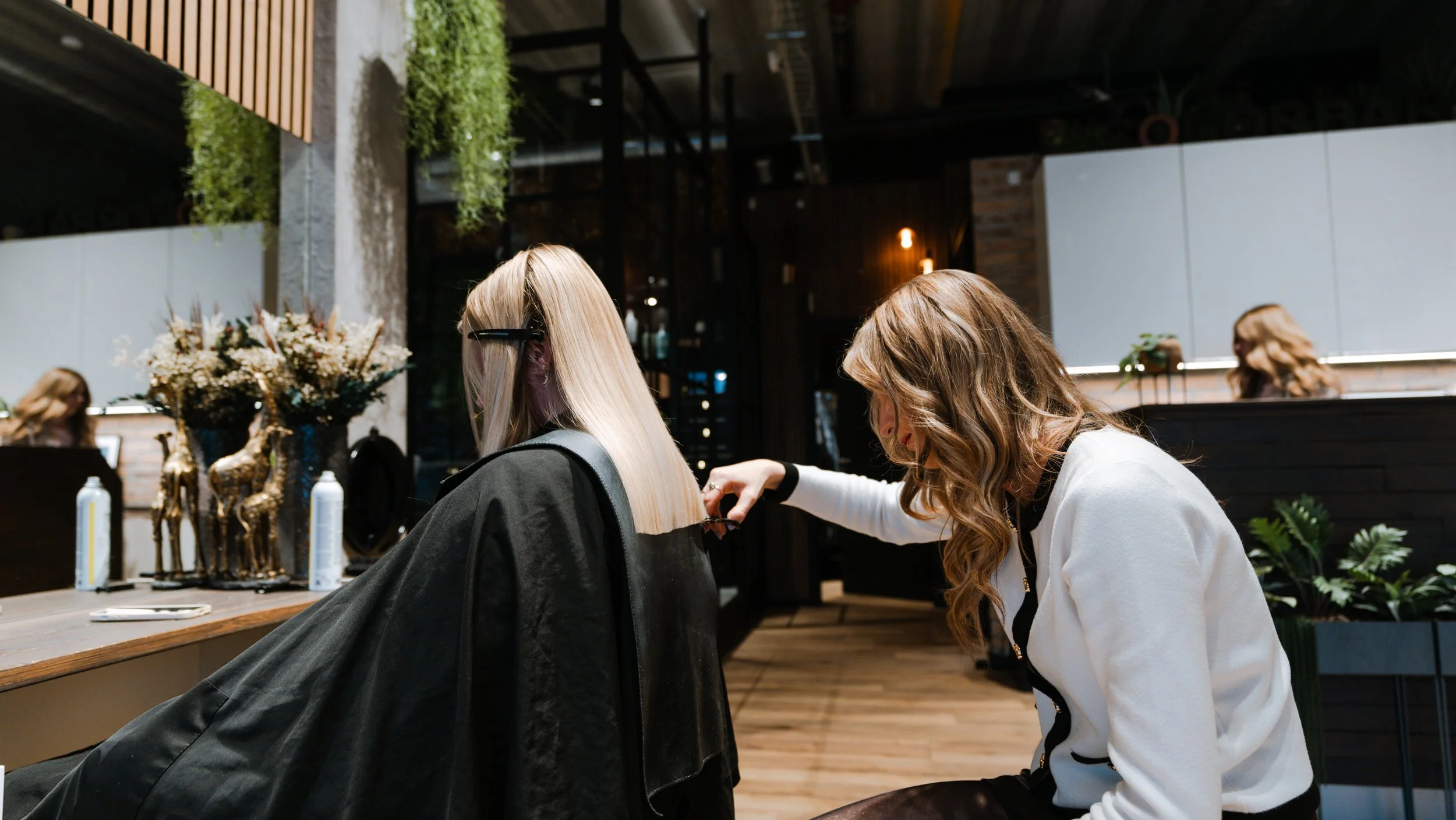 En frisør, der klipper en kundes blonde hår i en moderne frisørsalon med træ- og plantedekorationer.