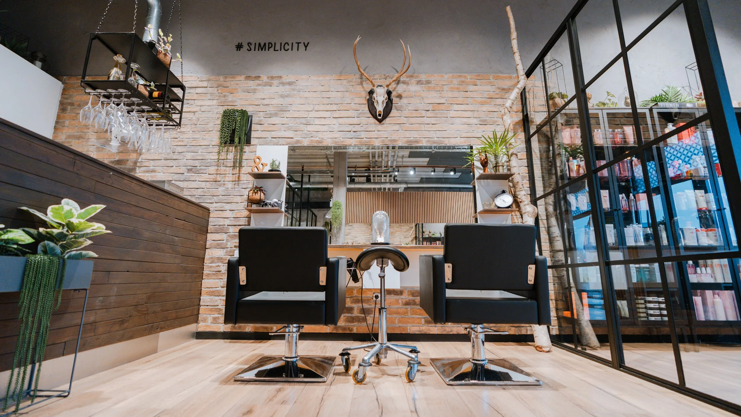 Indretning af en moderne frisørsalon med to sorte frisørstole foran en stor spejl, træpaneler, grønne planter, og en glasvæg med hårplejeprodukter. Der er en væg med murstenslook og et dyrehoved med gevirer.