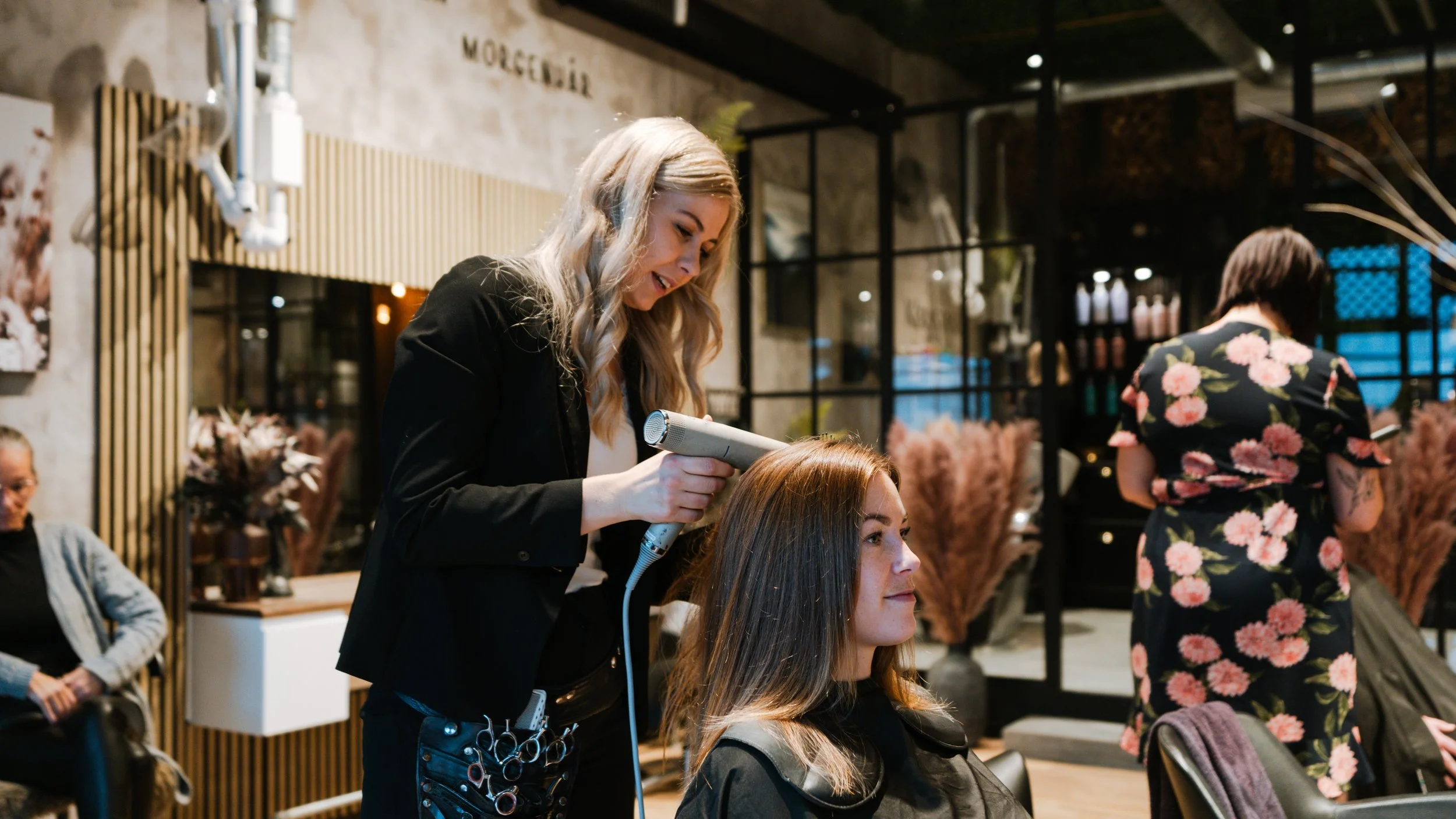 En frisør styling en kvindes hår med en føntørrer i en salon.