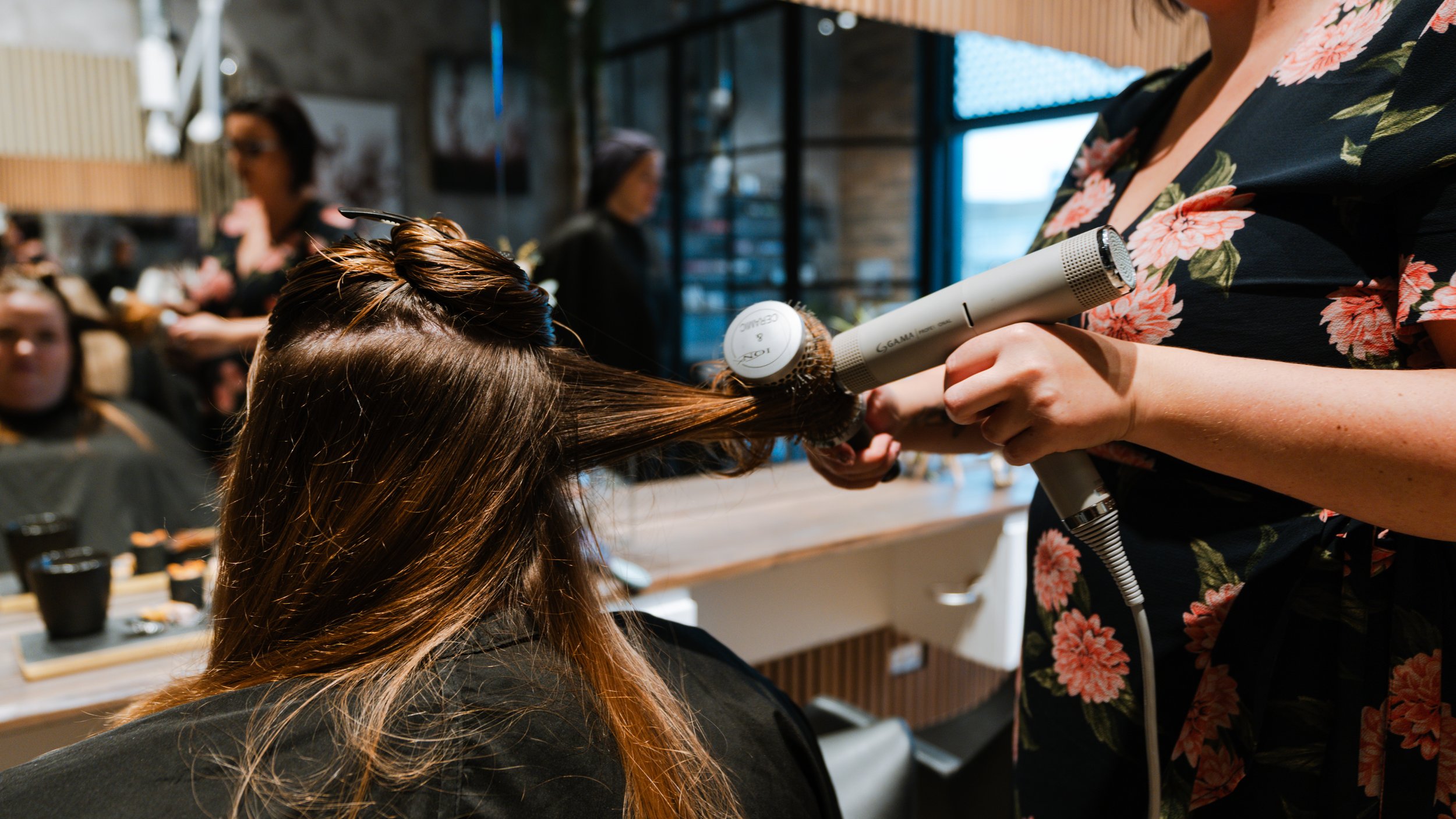 En frisør, der bruger en føntørre til at tørre en klients hår. Kunden sidder i en frisørstol, og der er andre kunder og frisører i baggrunden i salonen.